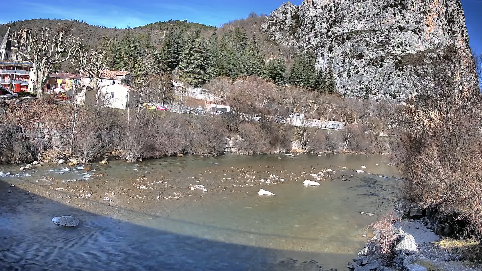 A slow-moving river flows past a village nestled at the base of a rocky cliff under a sunny sky.