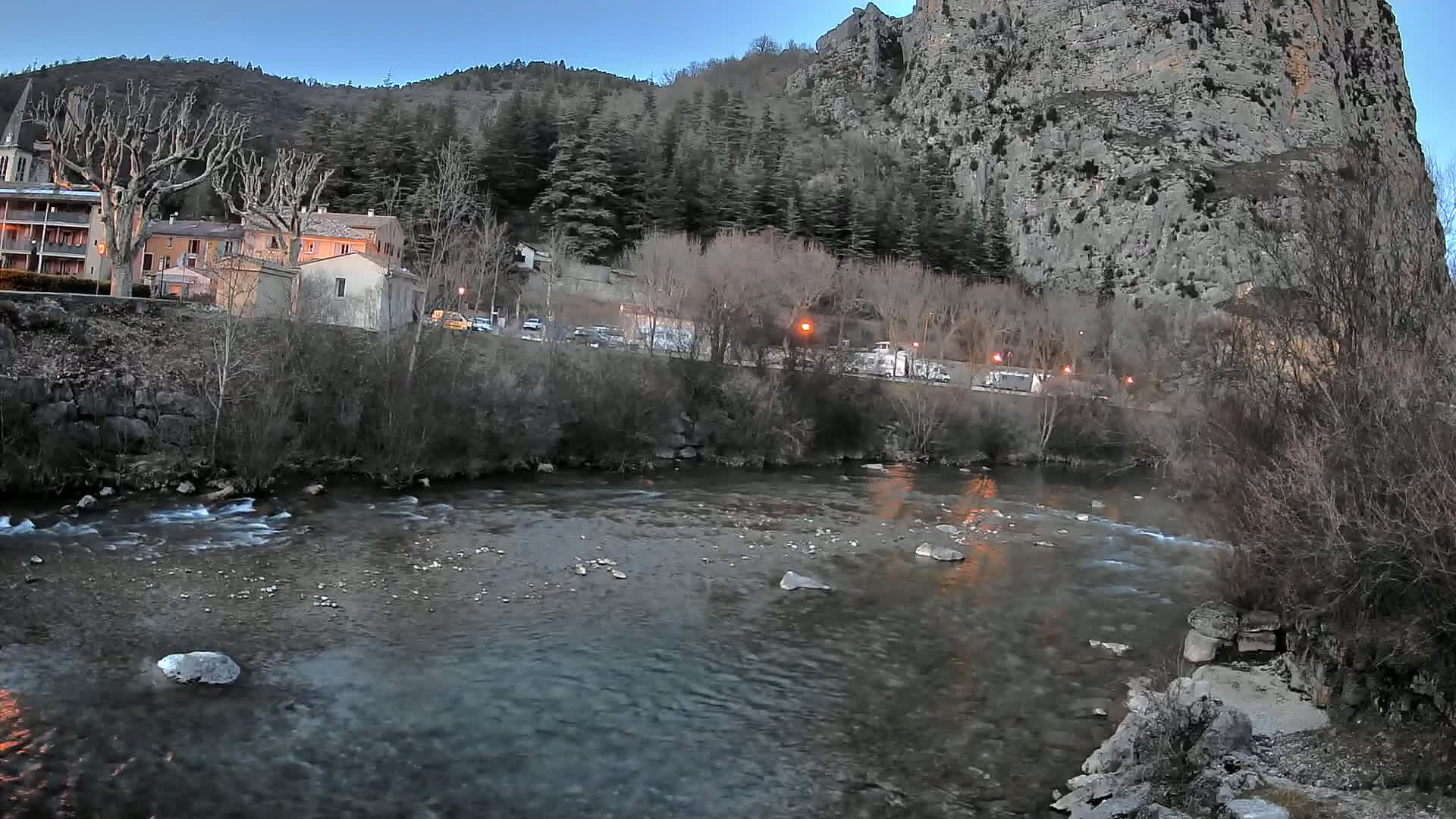 A river flows through a village nestled at the base of a large, rocky cliff under a twilight sky.