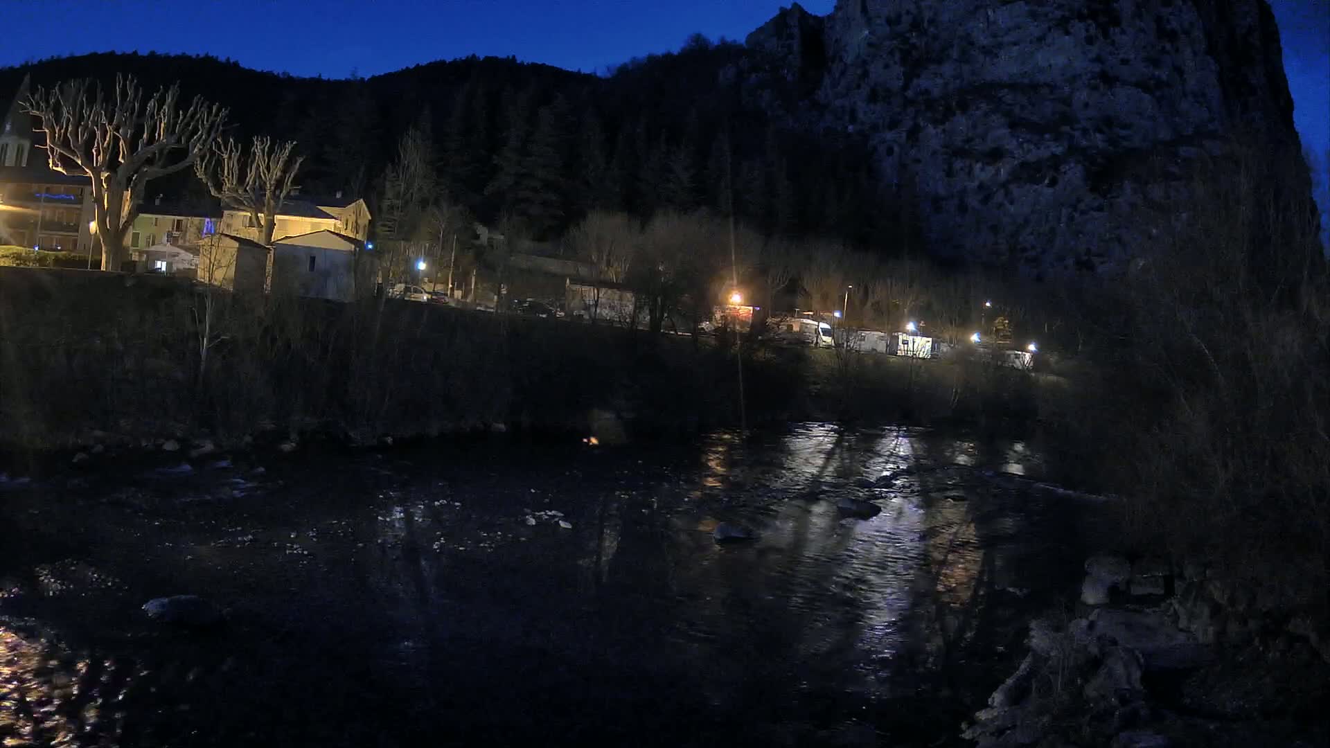 At night, a dark river flows past a small village nestled at the base of a large, dark cliff under a clear, dark blue sky.