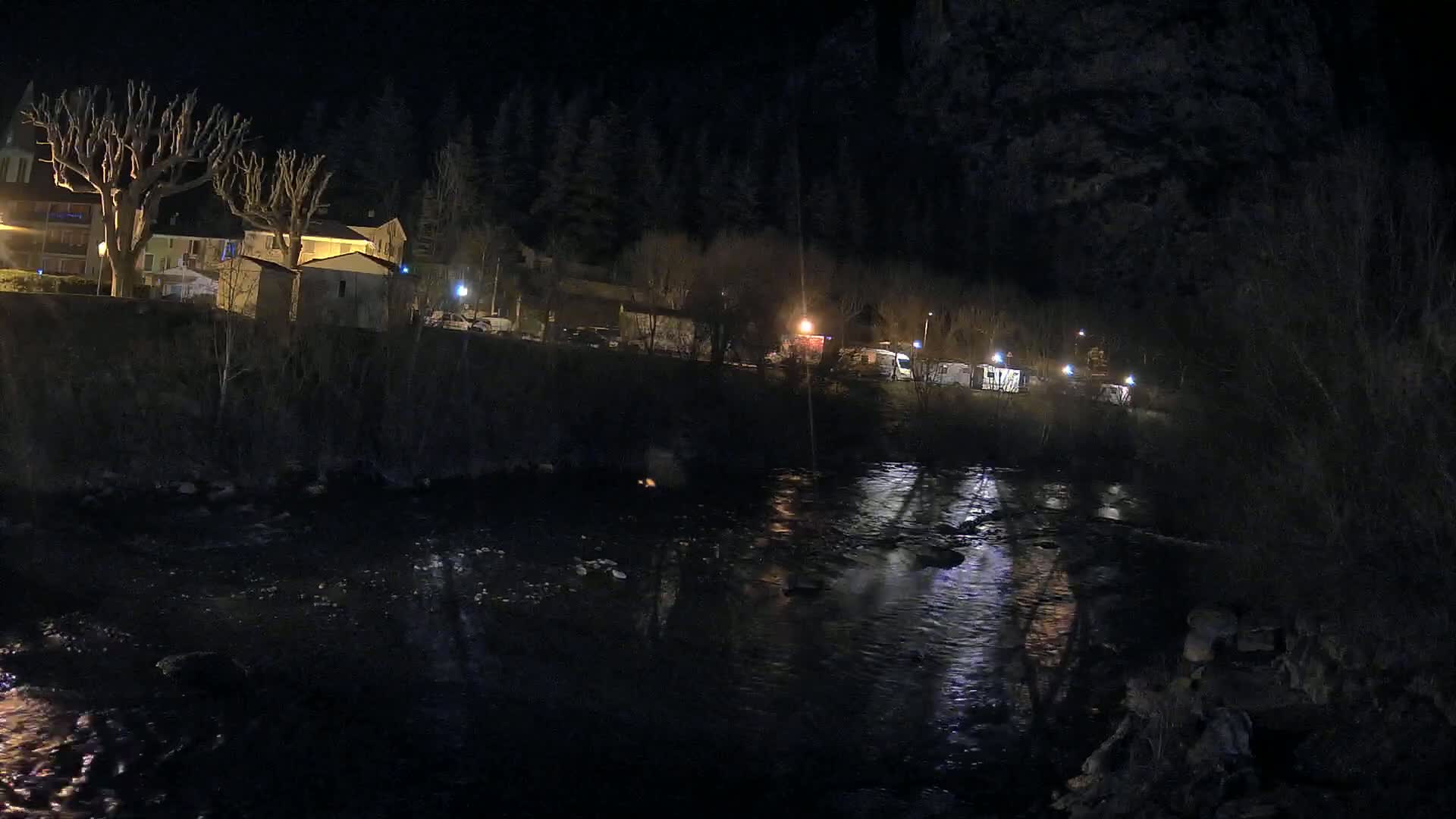 A dark, nighttime scene shows a river reflecting lights from buildings and vehicles along its bank, with bare trees and a wooded hillside in the background.