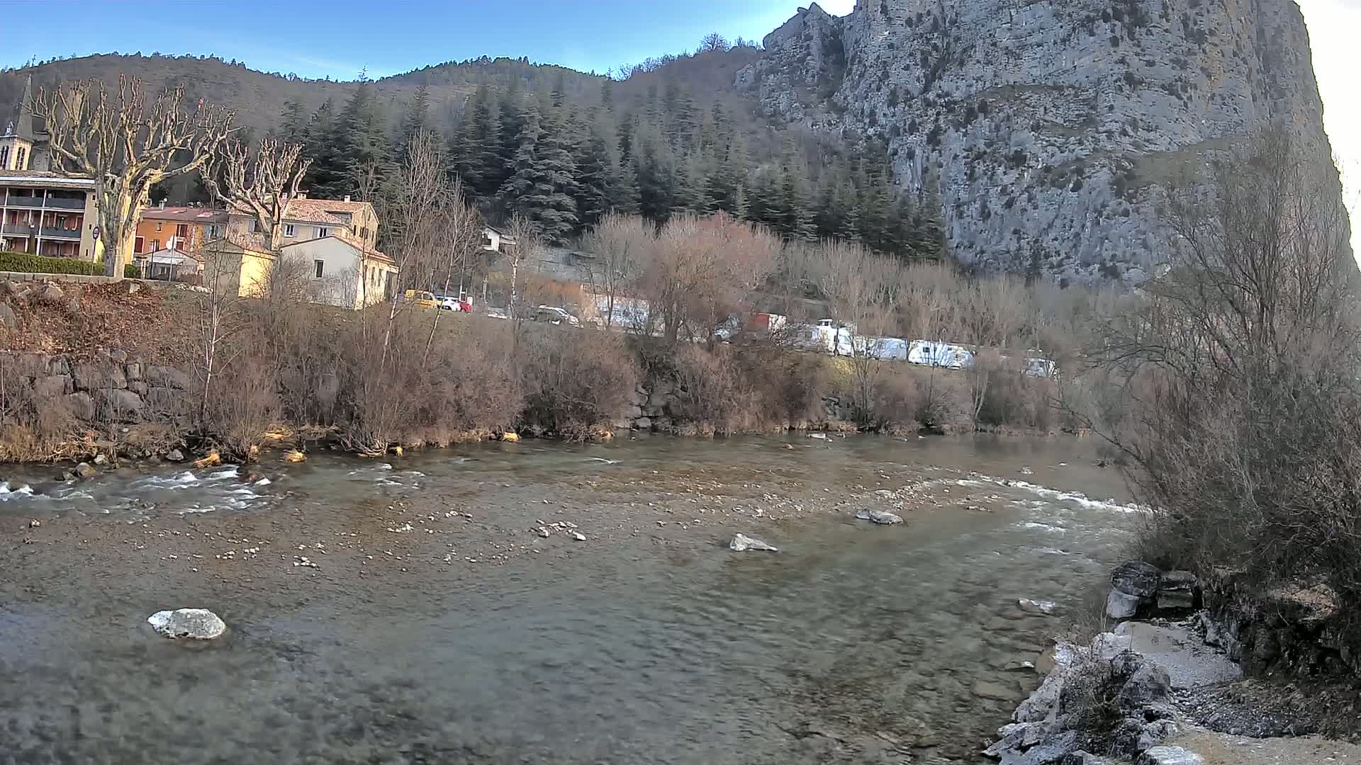 A clear, shallow river flows past a village nestled at the base of a steep, rocky cliff under a mostly clear sky.
