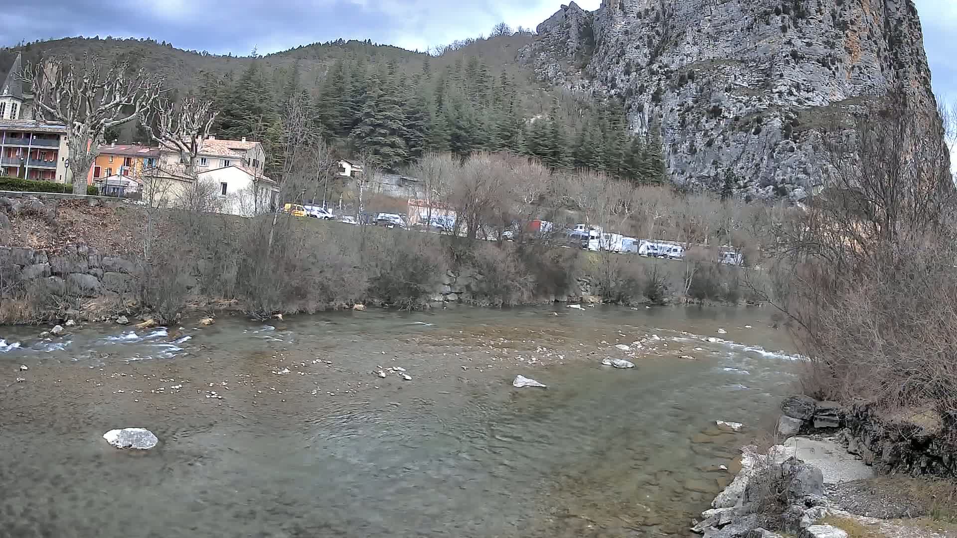 A shallow, rocky river flows past a village nestled at the base of a large, gray cliff under a cloudy sky.