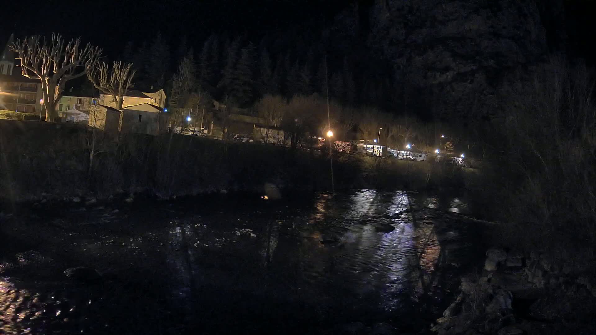 A dark river flows past a small village at night, illuminated by scattered streetlights against a backdrop of a dark forest.