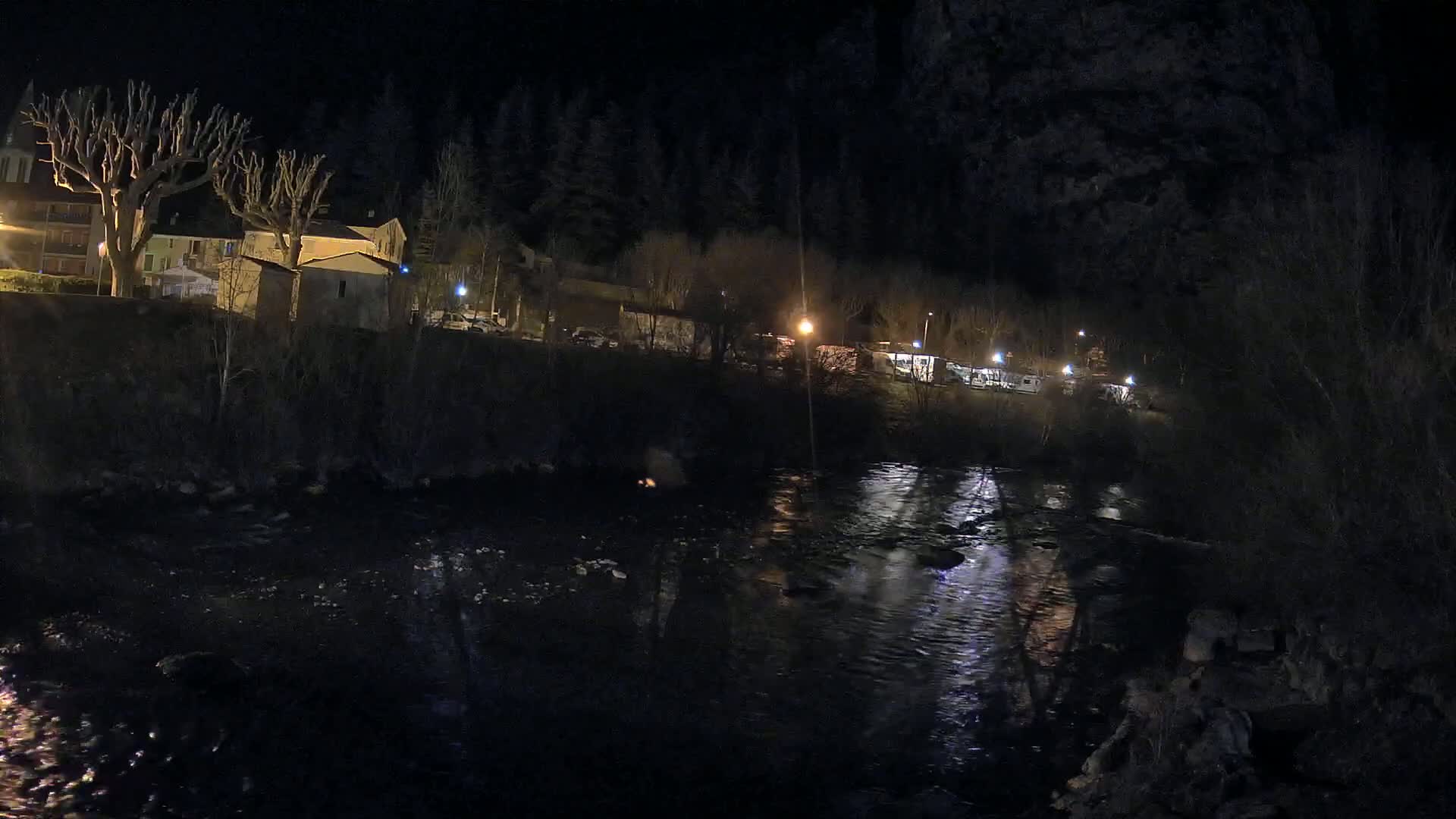 A dark, nighttime scene shows a river reflecting the lights of a small village nestled amongst dark trees on a hill.