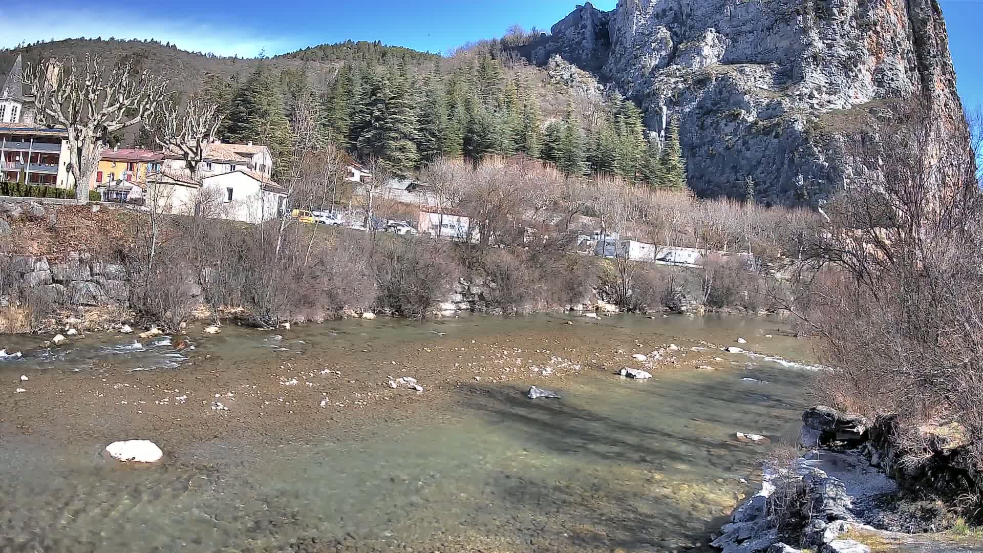 A clear, shallow river flows past a village nestled at the base of a large, rocky cliff under a sunny sky.