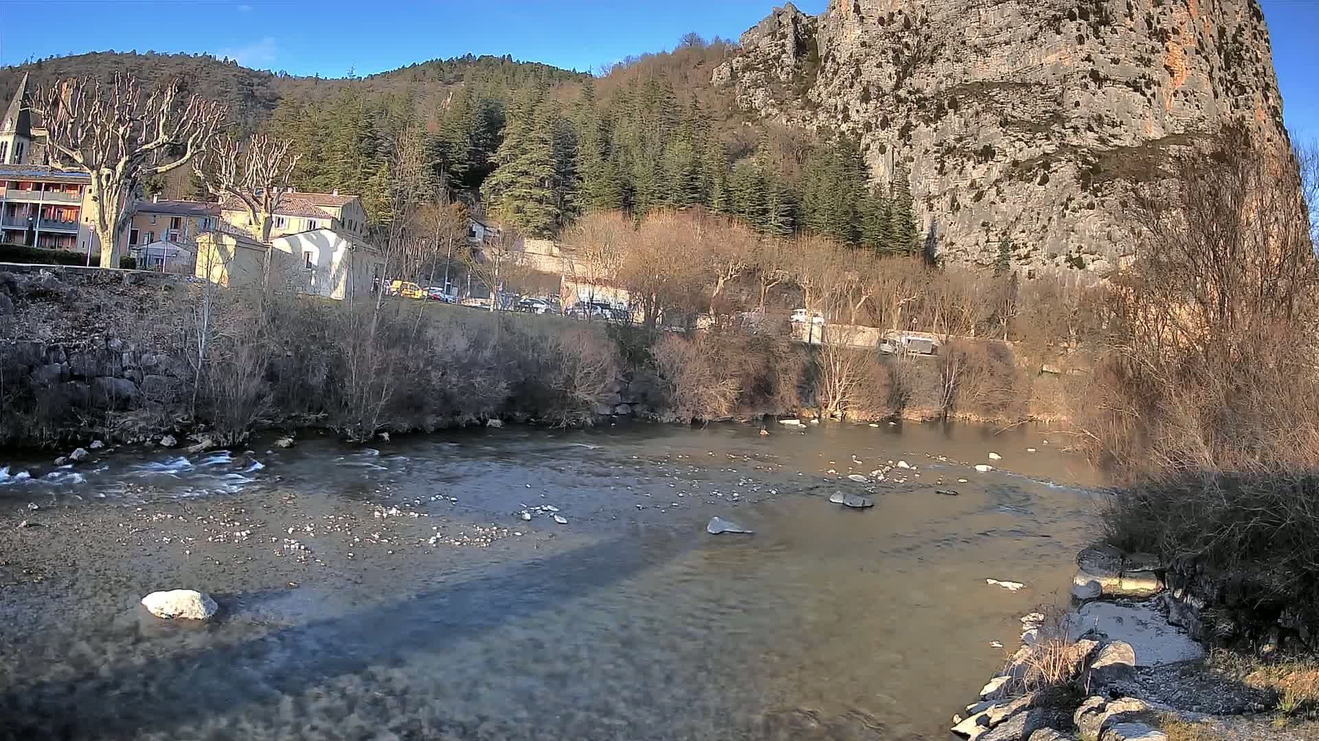 A river flows past a village nestled at the base of a large, rocky cliff face under a clear, sunny sky.