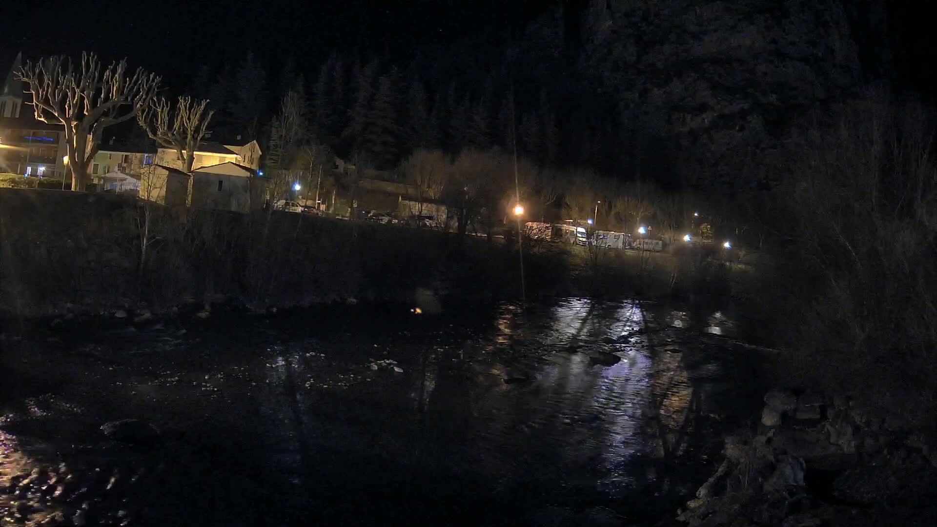 A river reflects the lights of a small village nestled beside a dark forest at night.