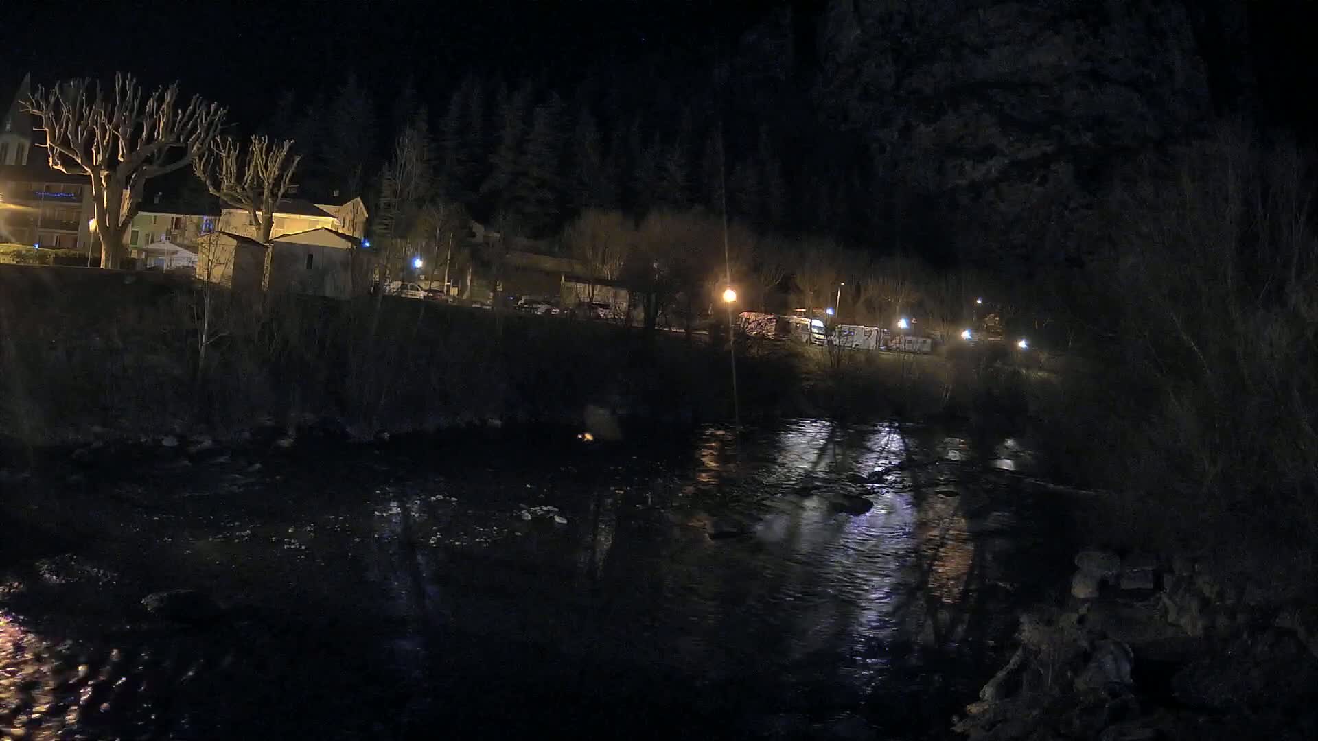 A dark, nighttime scene shows a river reflecting the lights of a small village nestled beside a dark forest.