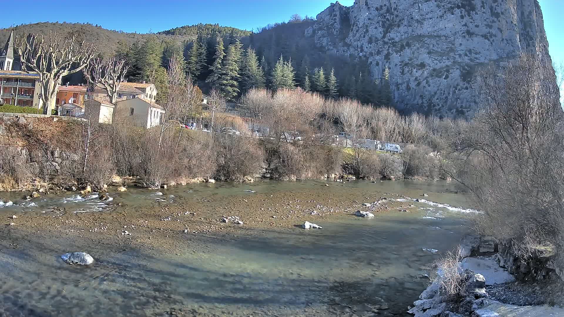 A shallow, rocky river flows past a village nestled at the base of a large, gray cliff under a clear blue sky.