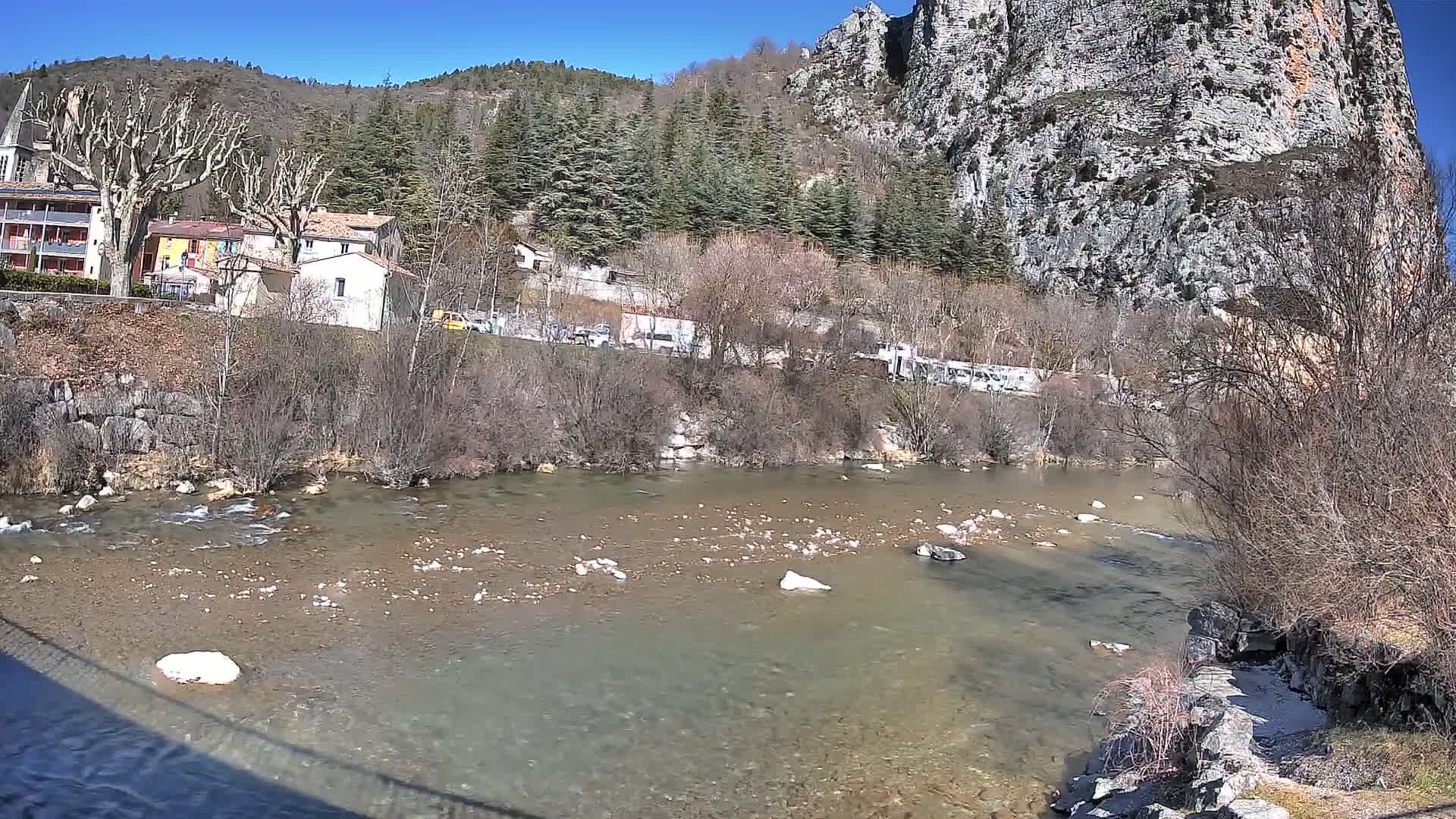 A clear, shallow river flows past a village nestled at the base of a rocky cliff under a sunny sky.