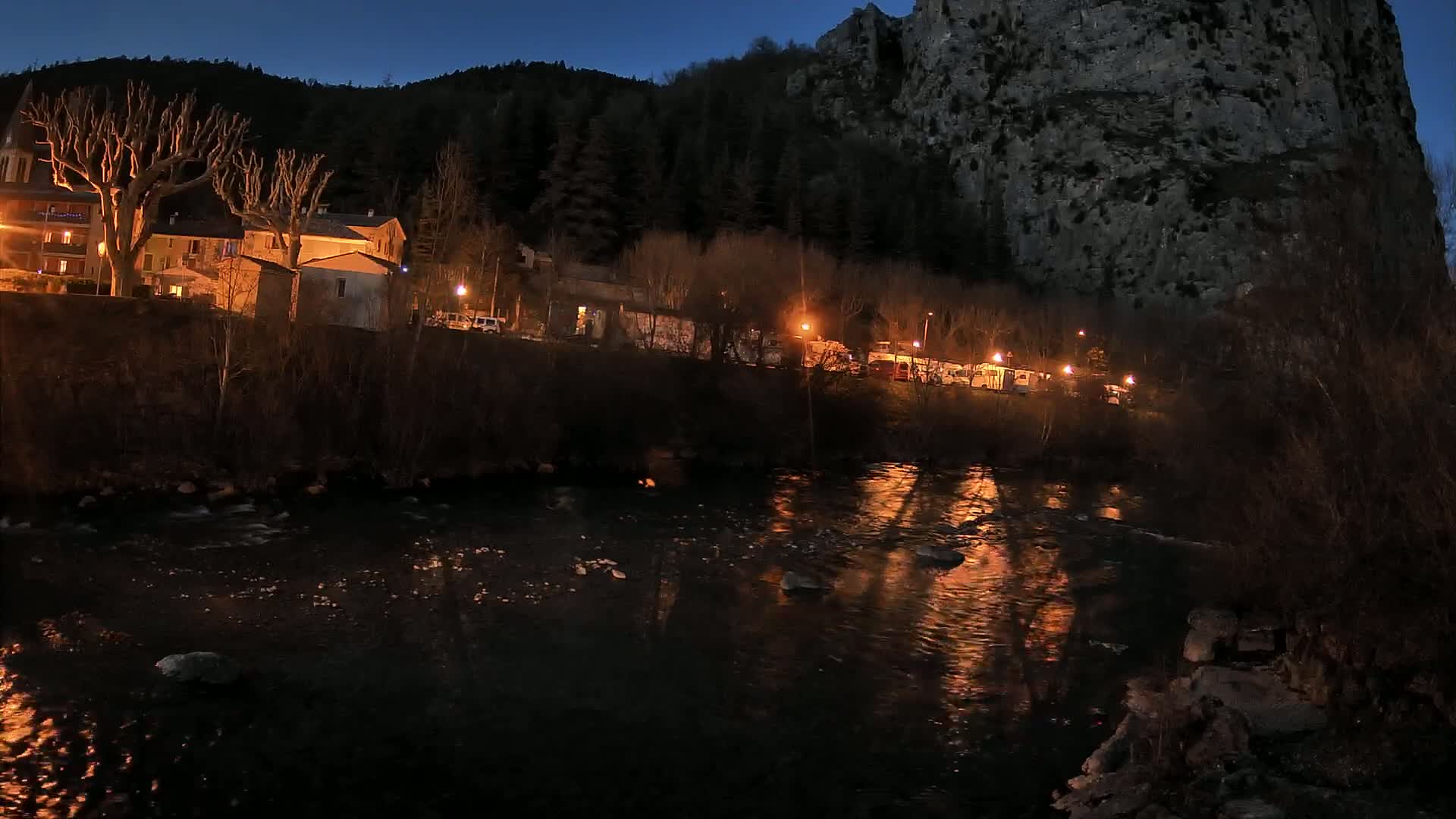 A river reflects the lights of a small village nestled at the base of a dark, rocky cliff under a twilight sky.