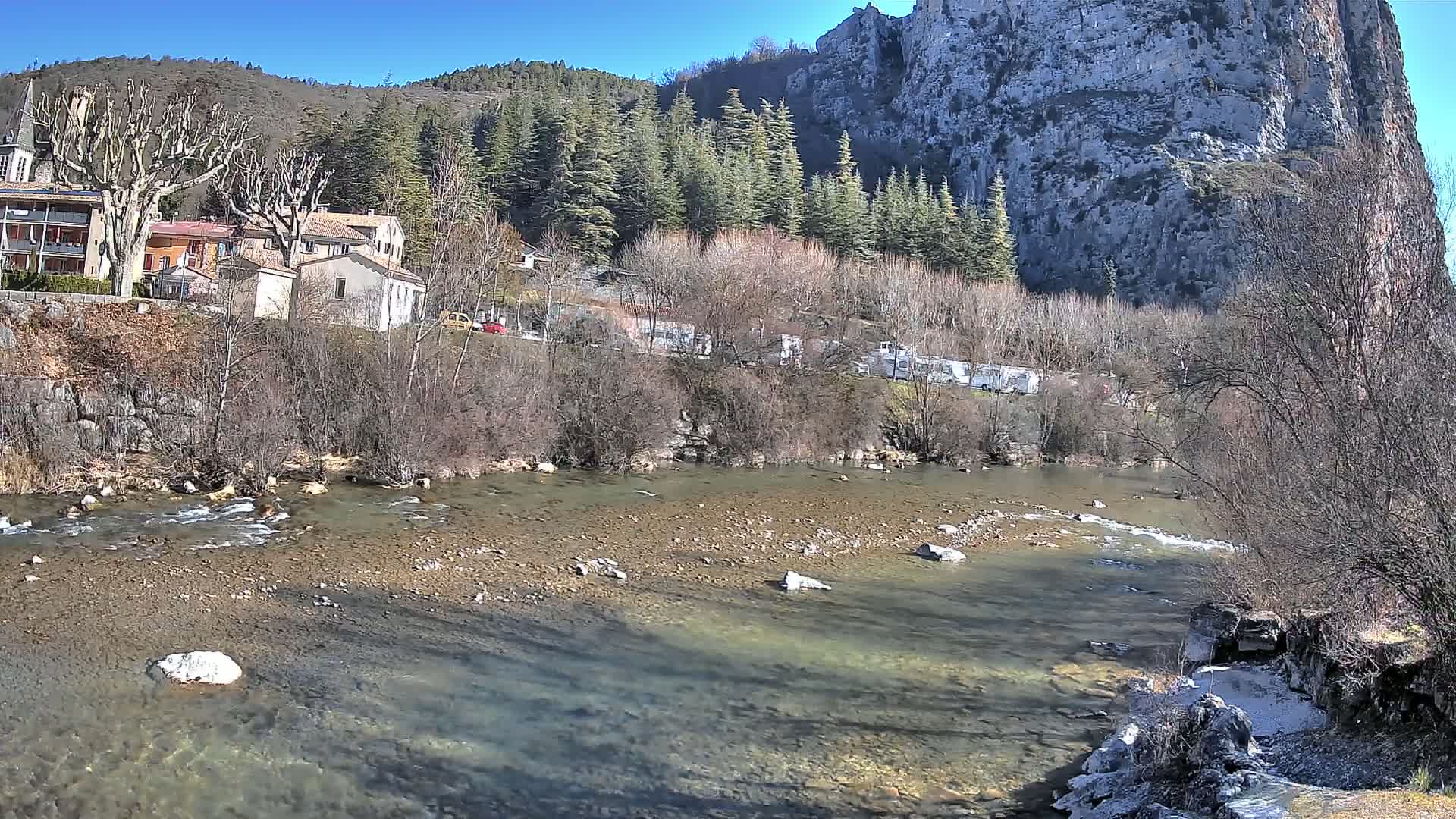 A clear river flows past a village nestled at the base of a rocky cliff, under a sunny sky.