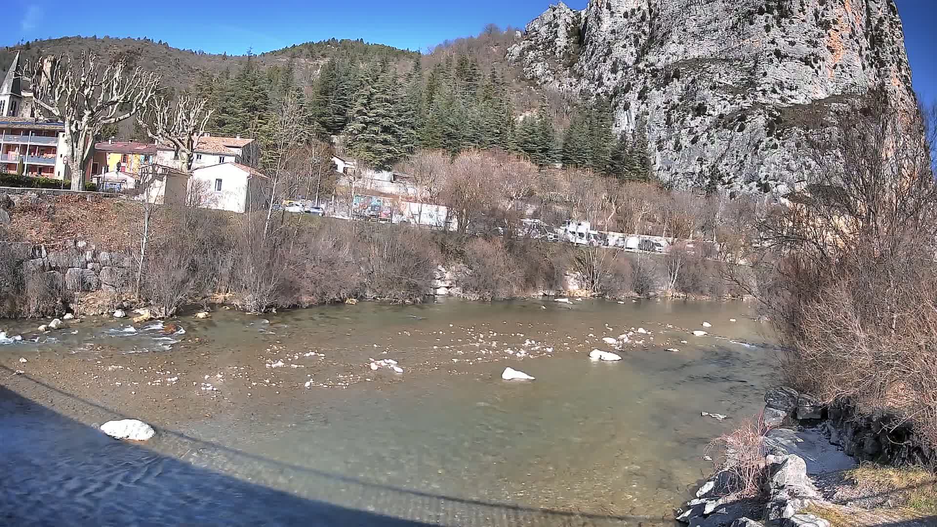A clear river flows past a small village nestled beneath a steep, rocky cliffside on a sunny day.