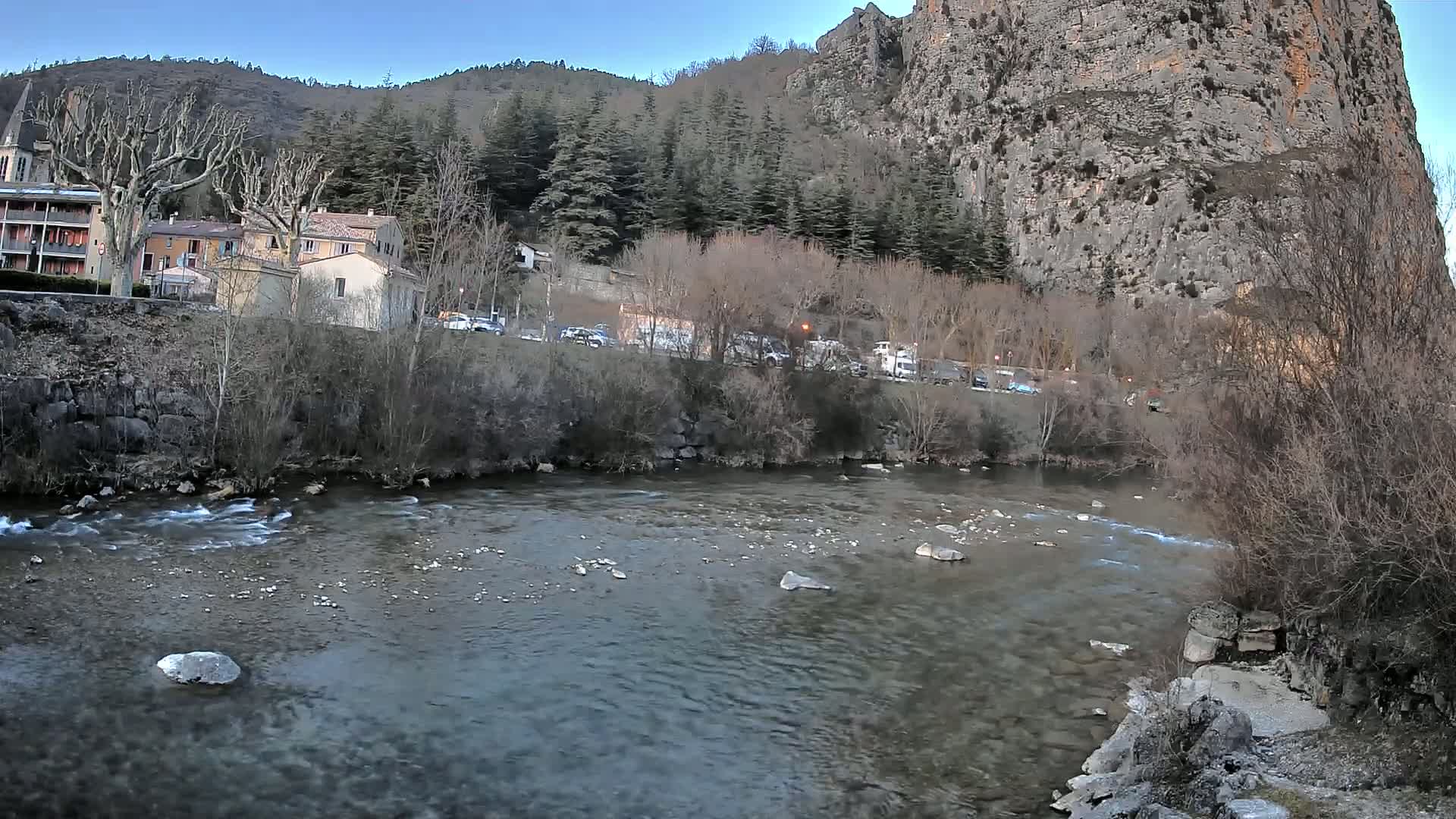 A clear river flows past a village nestled at the base of a large, rocky cliff under a twilight sky.