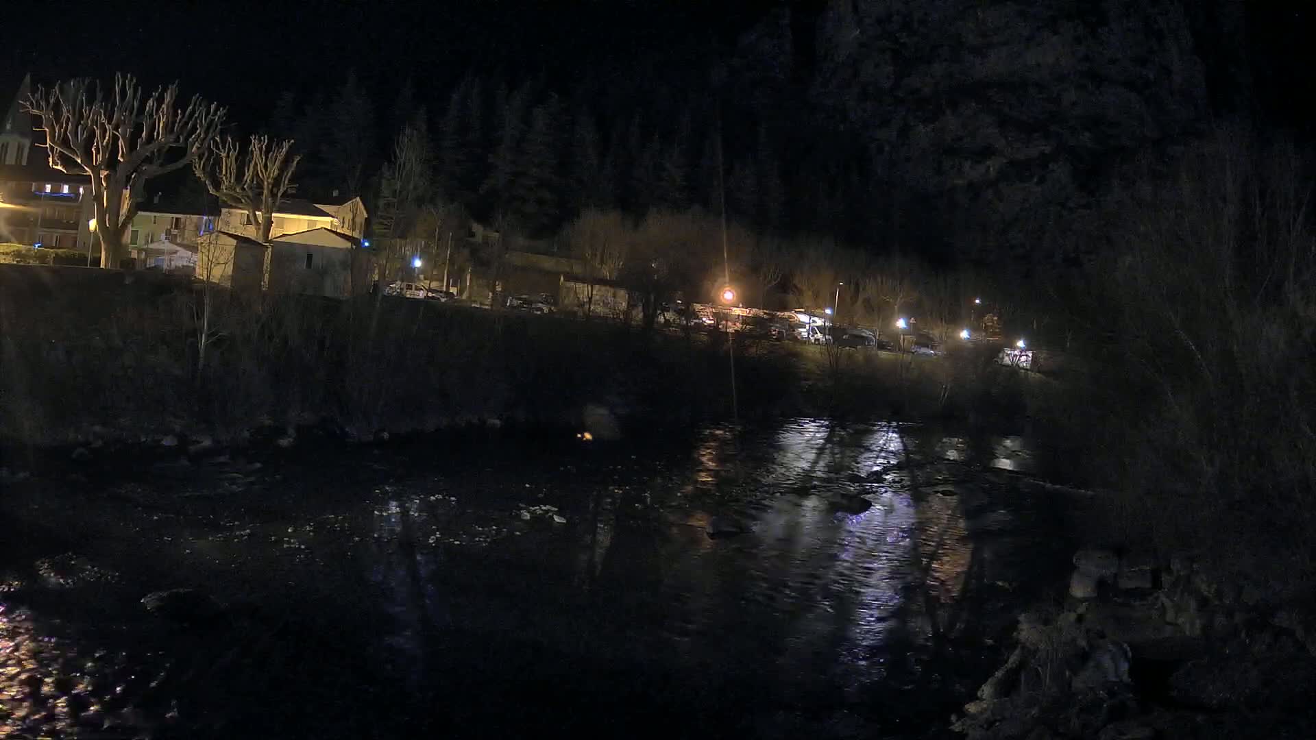 A dark river flows past a nighttime village nestled beside a dark forest under a clear night sky.