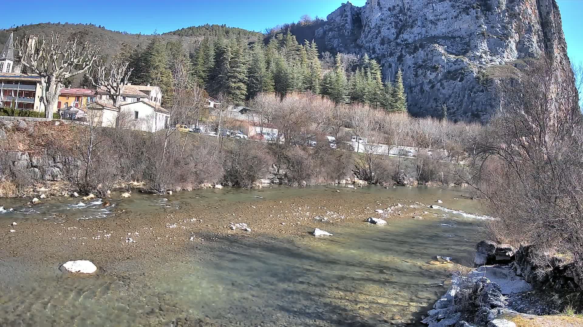 A clear river flows past a small village nestled at the base of a large, rocky cliff under a sunny sky.