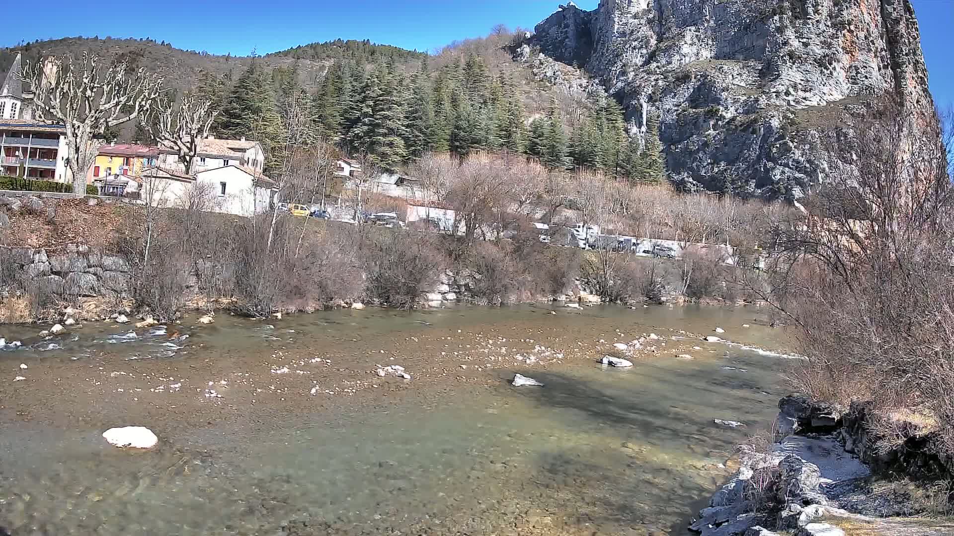 A clear river flows past a village nestled at the base of a large, rocky cliff under a bright, sunny sky.