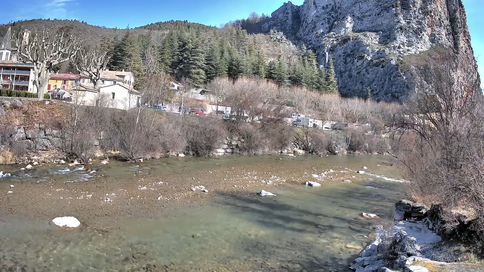 A clear, shallow river flows past a village nestled at the base of a large, rocky cliff under a sunny sky.