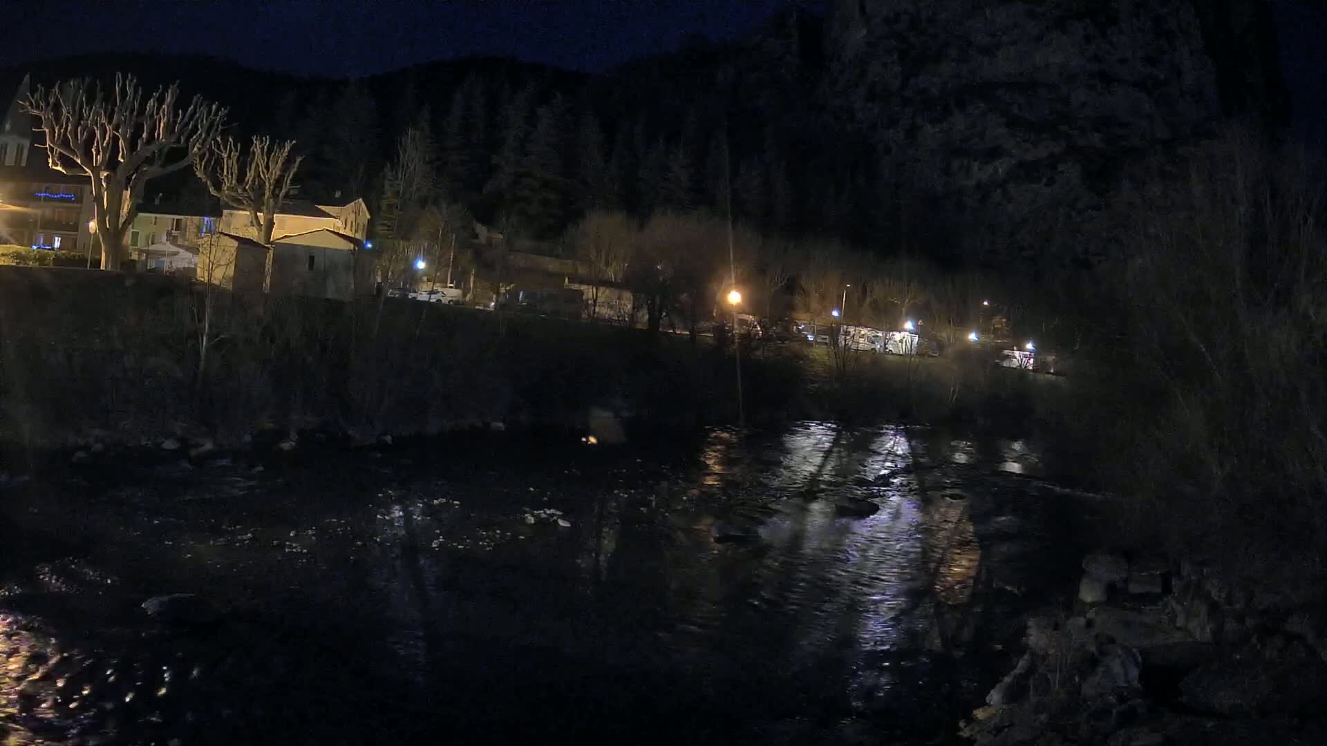 A dark, nighttime scene shows a river flowing past a small village nestled against a hillside under a clear, starless sky.