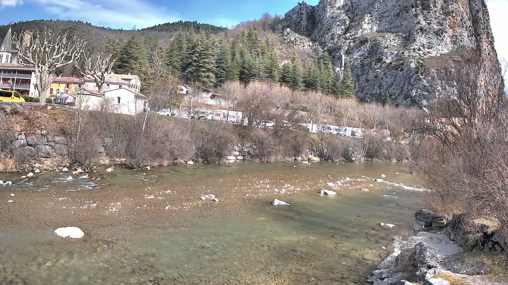 A clear river flows past a village nestled at the base of a large, rocky cliff under a sunny sky.