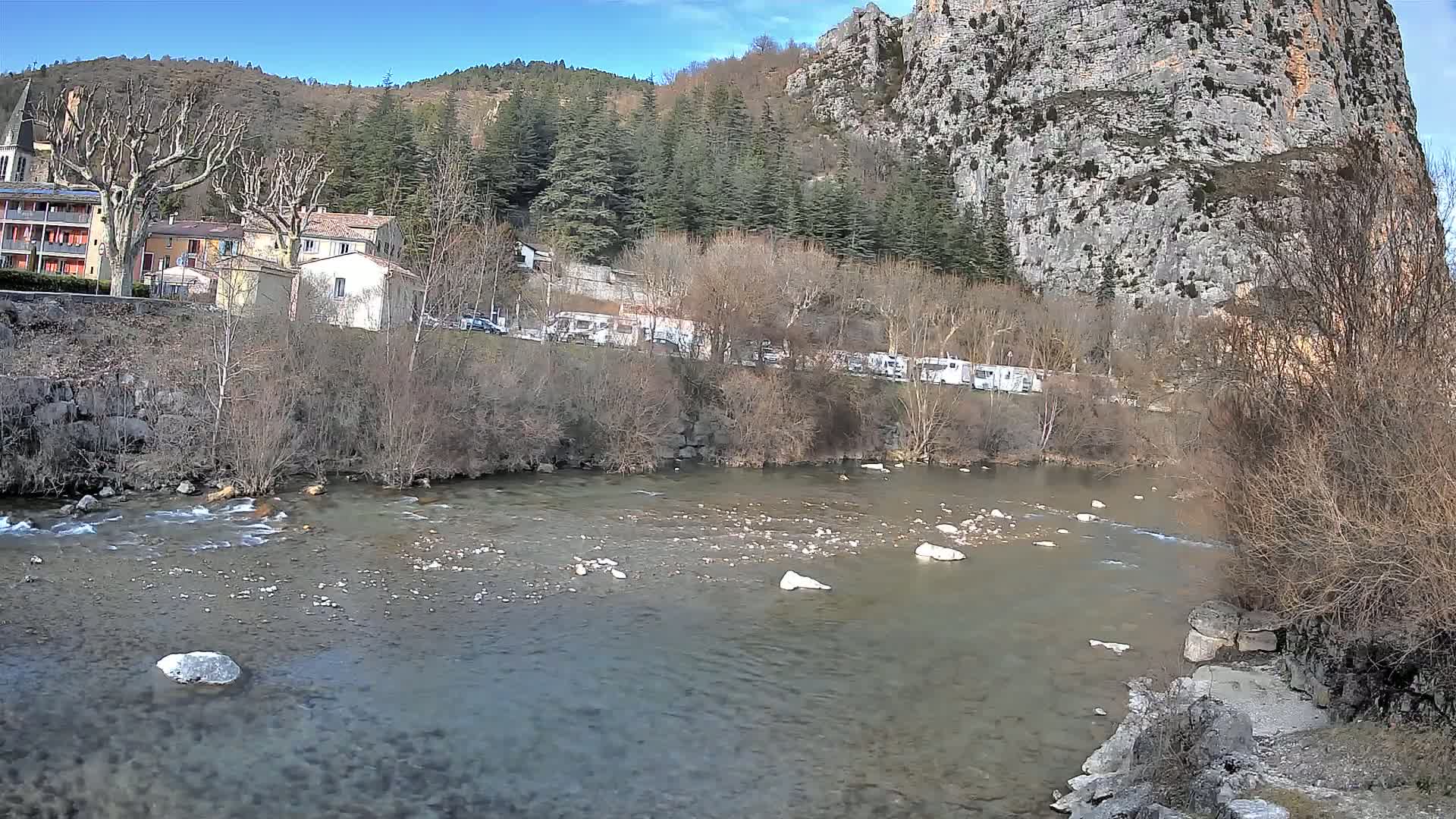 A clear river flows past a village nestled at the base of a large, rocky cliff under a bright, sunny sky.