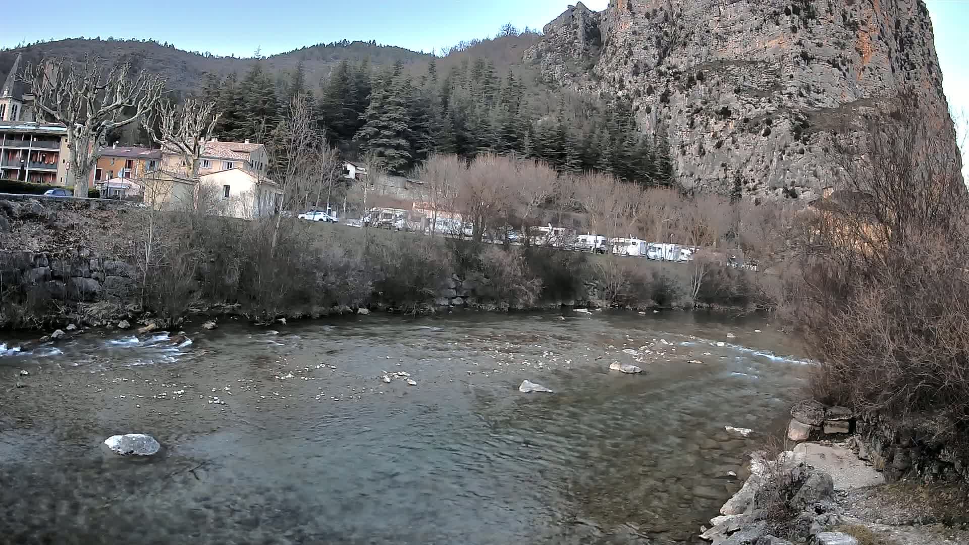 A clear river flows past a village nestled beside a large rock face and a forest under a mostly clear sky.