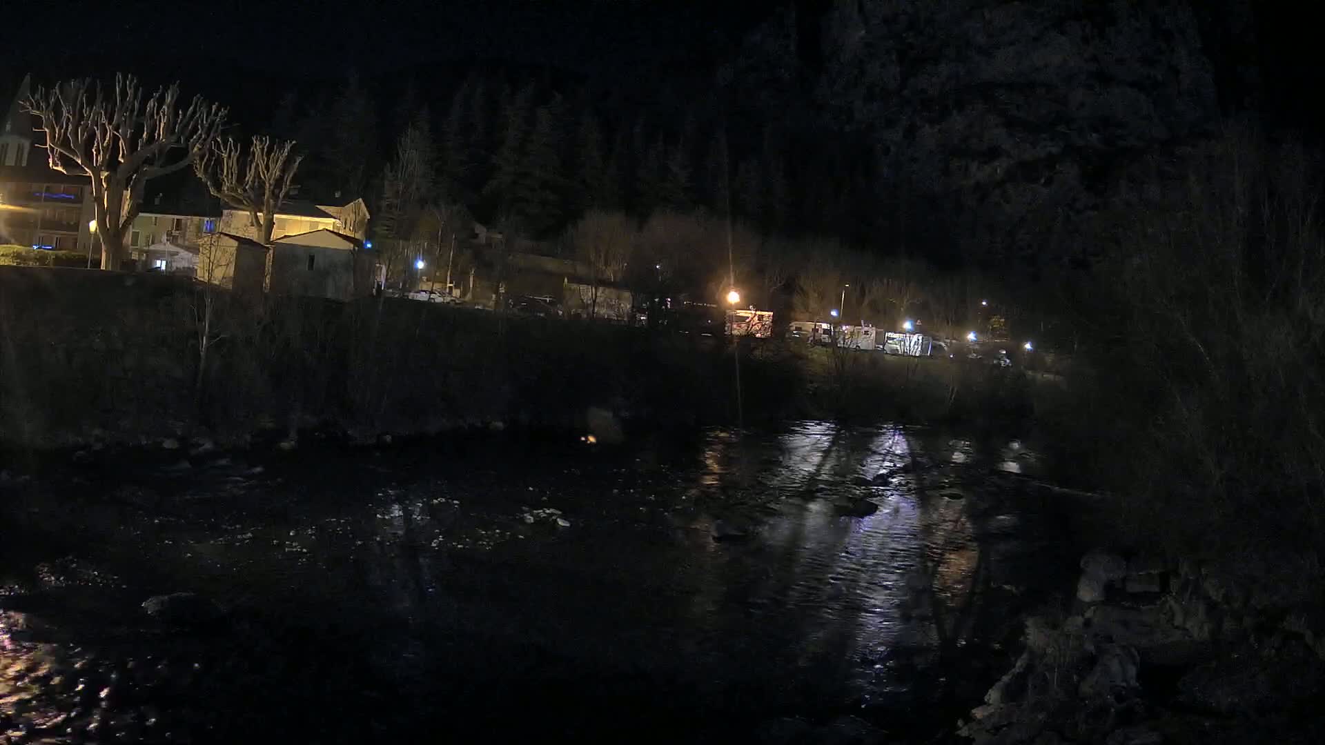 A dark river reflects the lights of a village at night, with bare trees and buildings lining the riverbank.
