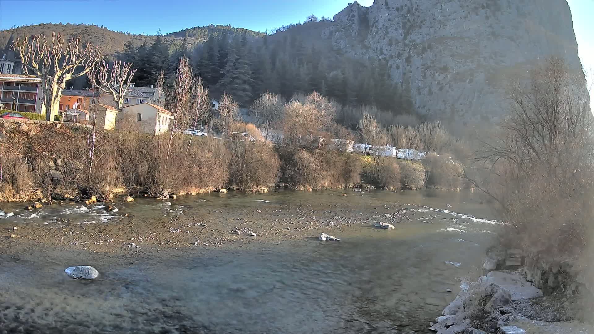 A slow-moving river flows past a village nestled at the base of a large, rocky cliff under a clear, sunny sky.