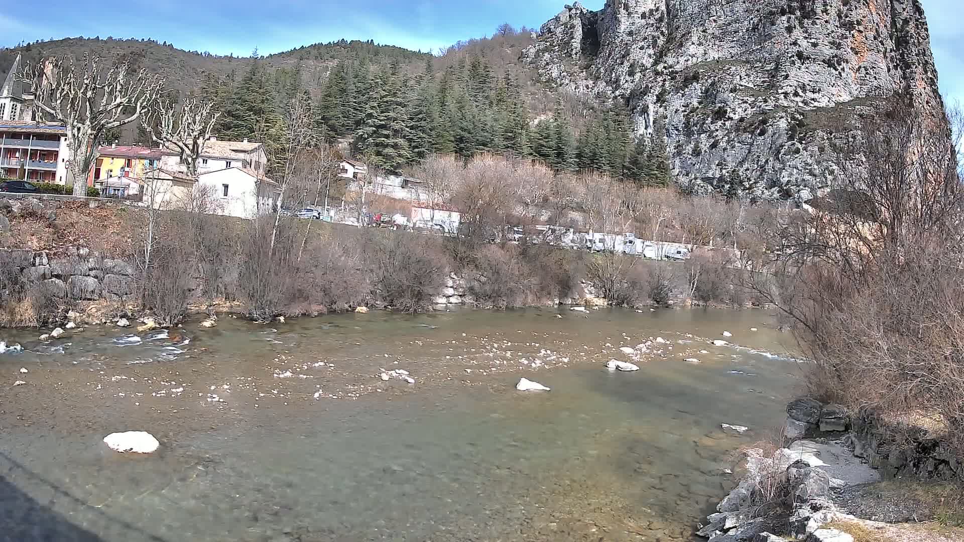 A clear river flows past a village nestled at the base of a large, rocky cliff under a sunny sky.