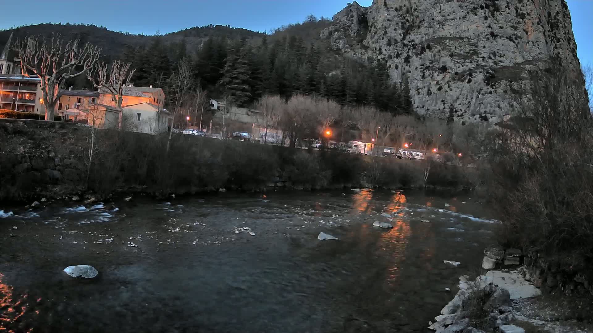 A river flows past a village nestled at the base of a large, rocky cliff under a twilight sky.