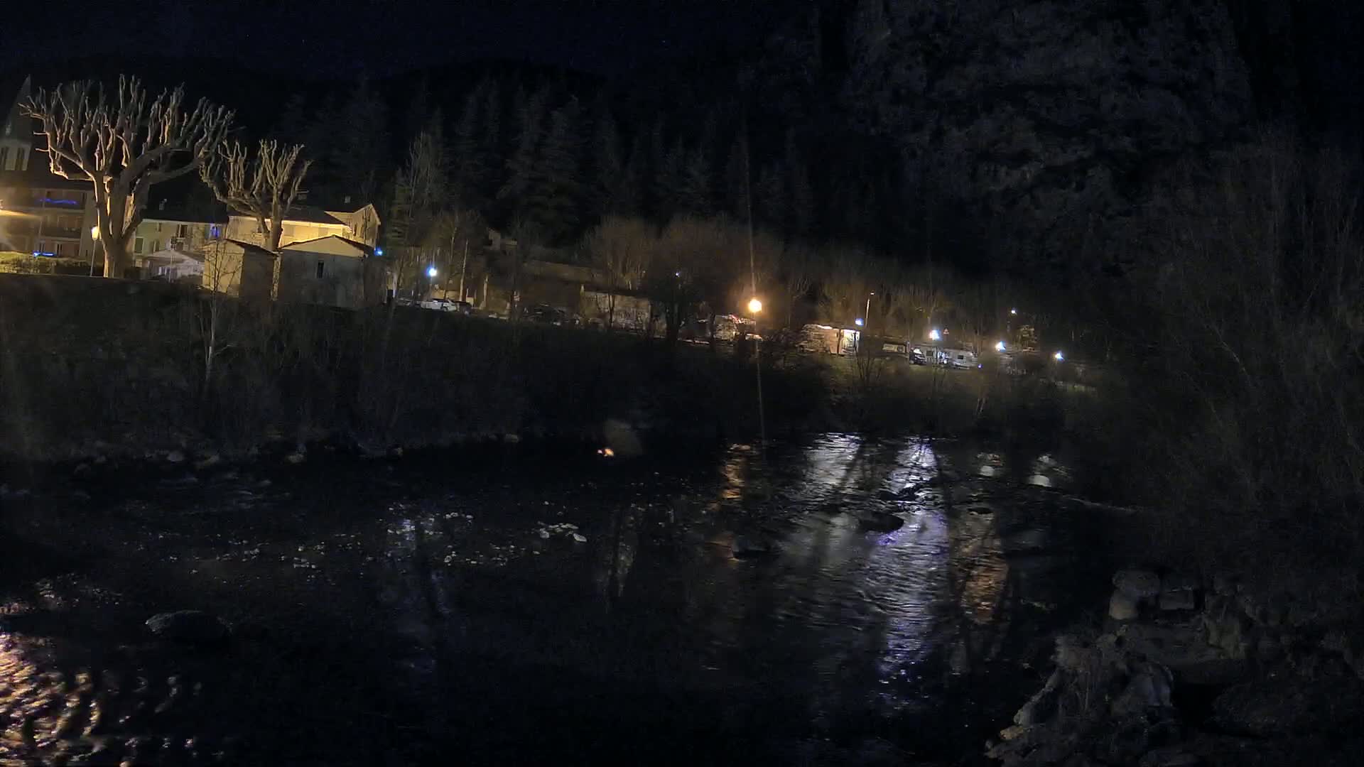 A dark, flowing river reflects the lights of a small village nestled at the foot of a wooded hill at night.