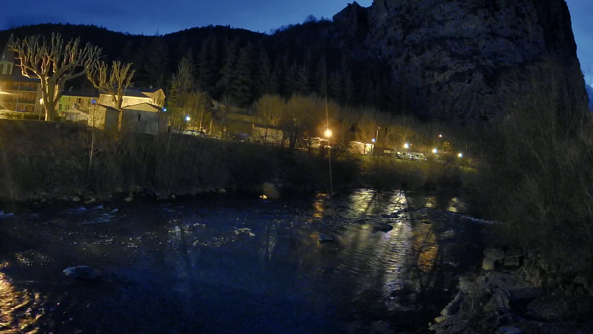 A dark, calm river flows past a small village nestled at the base of a large, dark cliff under a twilight sky.