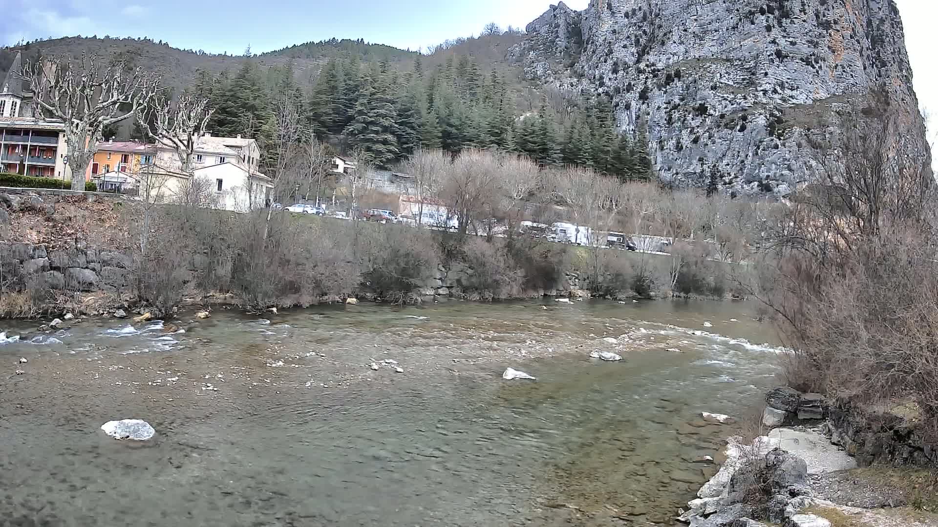 A shallow, rocky river flows past a village nestled beside a steep, grey cliff under a partly cloudy sky.