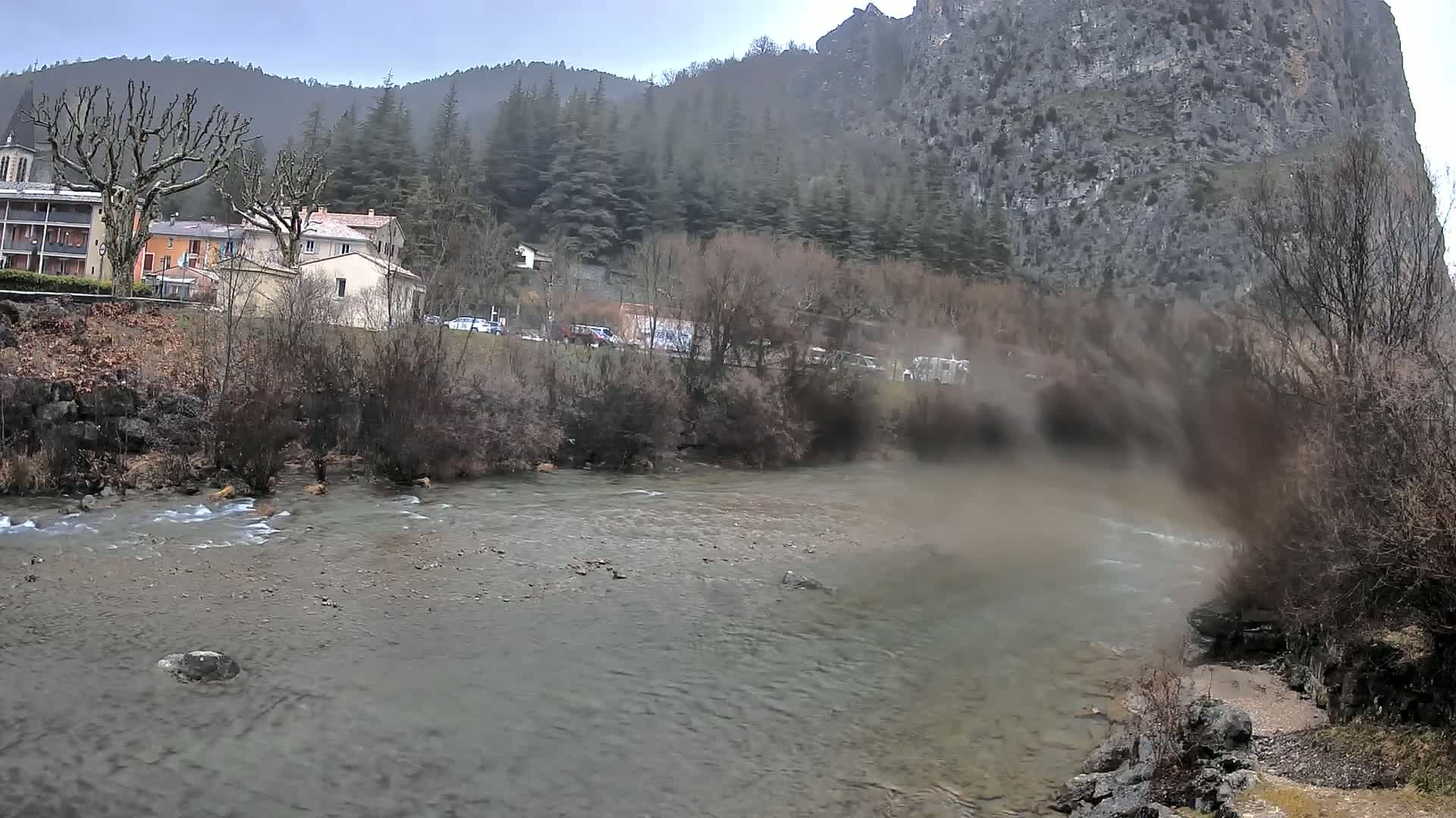 A river flows gently past a small village nestled at the base of a large, grey cliff under an overcast sky.