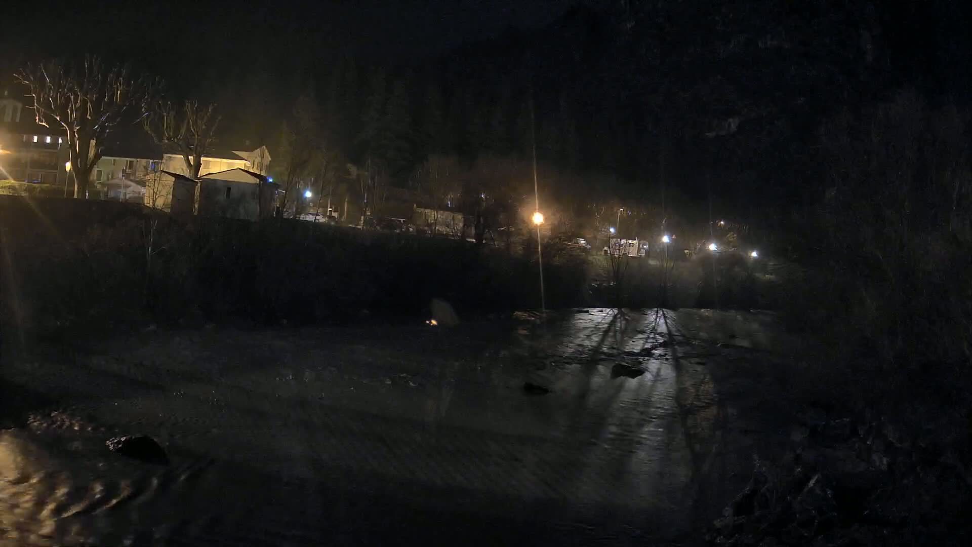 A dark, nighttime scene shows a river flowing past buildings and trees under dim, artificial light.