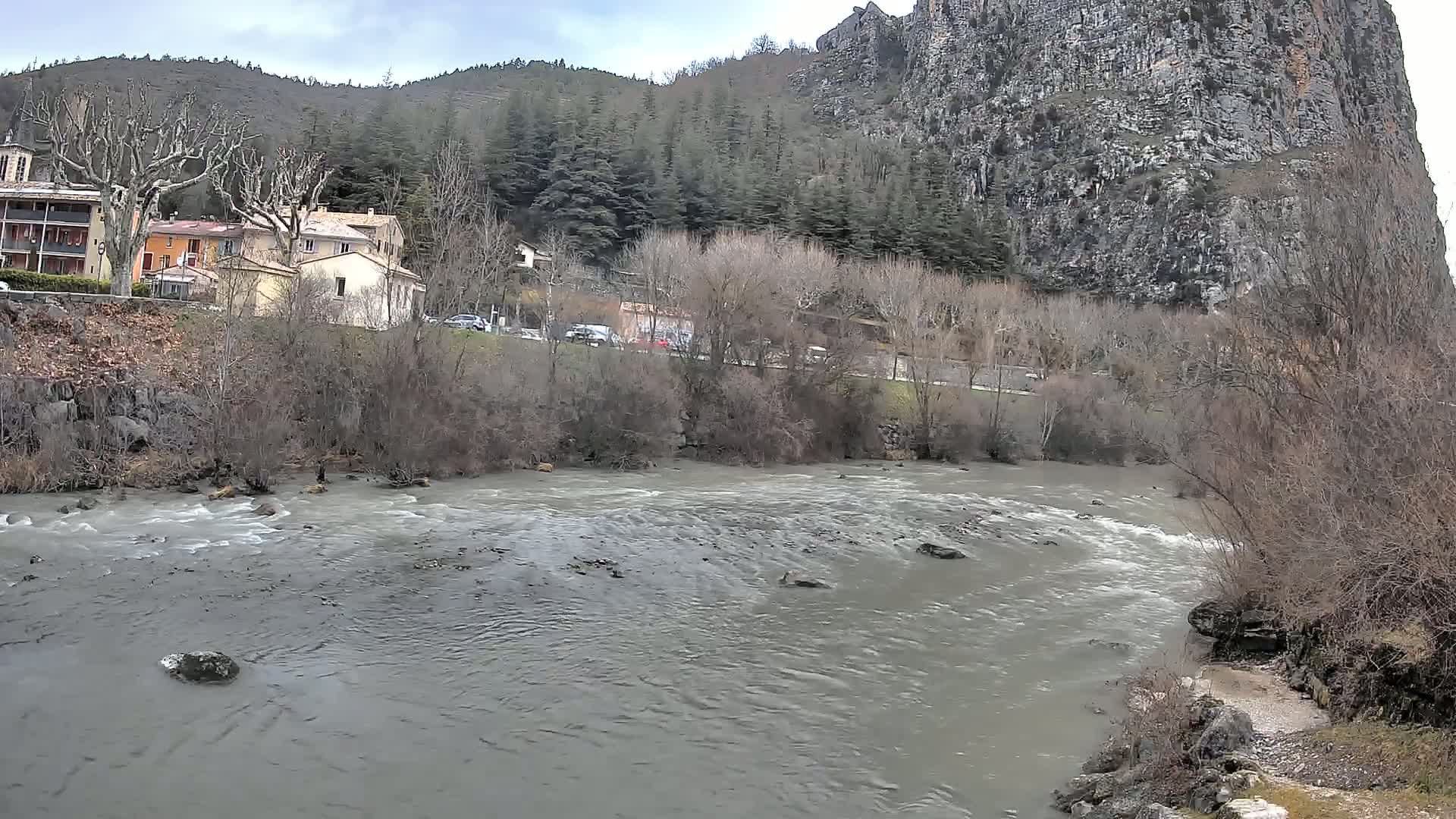 A murky river flows past a village nestled beside a steep, rocky cliff under an overcast sky.