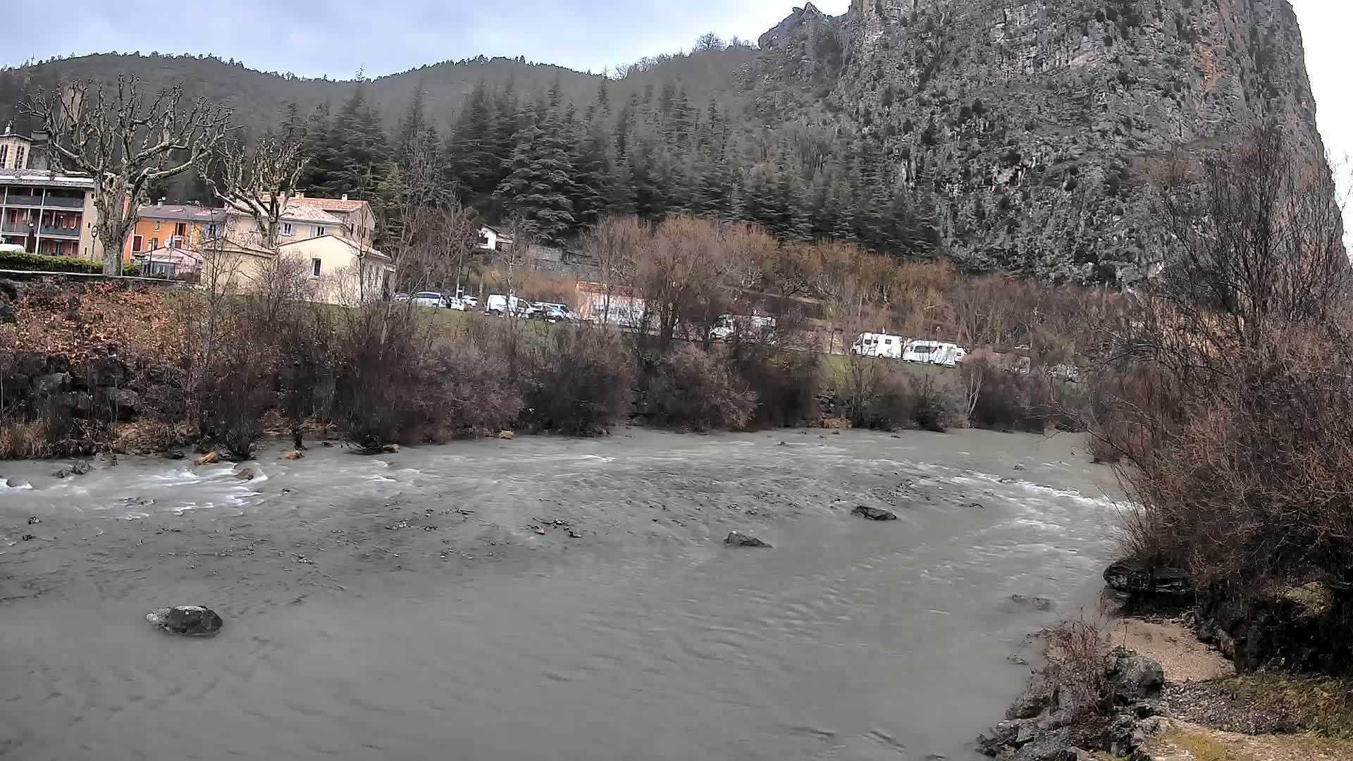 A murky river flows past a village nestled beside a steep, rocky mountain under an overcast sky.