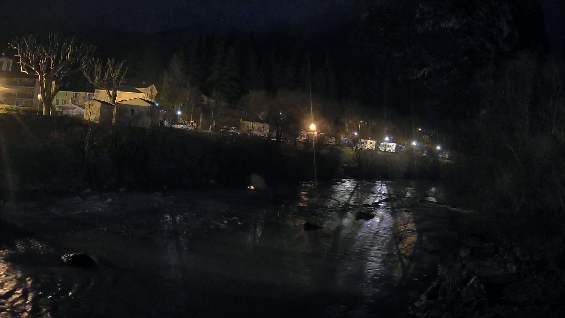 A dark, flowing river at night reflects the sparse lights of buildings and vehicles on the opposite bank, under a clear sky.