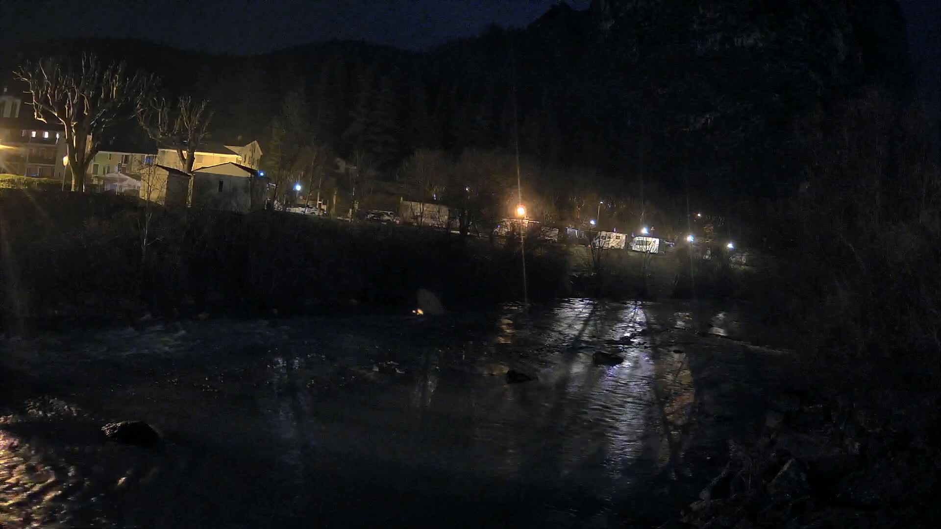 A dark, nighttime scene shows a river reflecting lights from buildings and vehicles along its bank, nestled against a hillside under a clear, moonless sky.