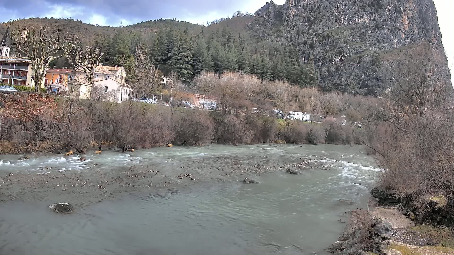 A cloudy day reveals a flowing river in front of a village nestled at the base of a large, rocky cliff and a forested hillside.