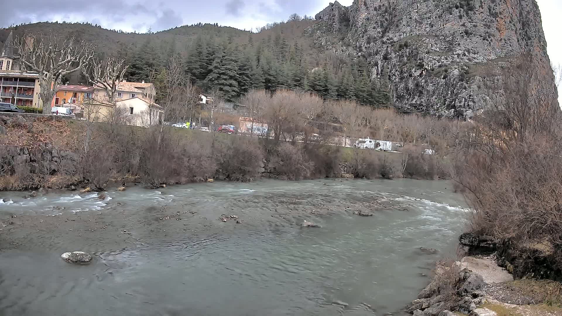 A river flows past a village nestled at the base of a large, rocky cliff under a cloudy sky.