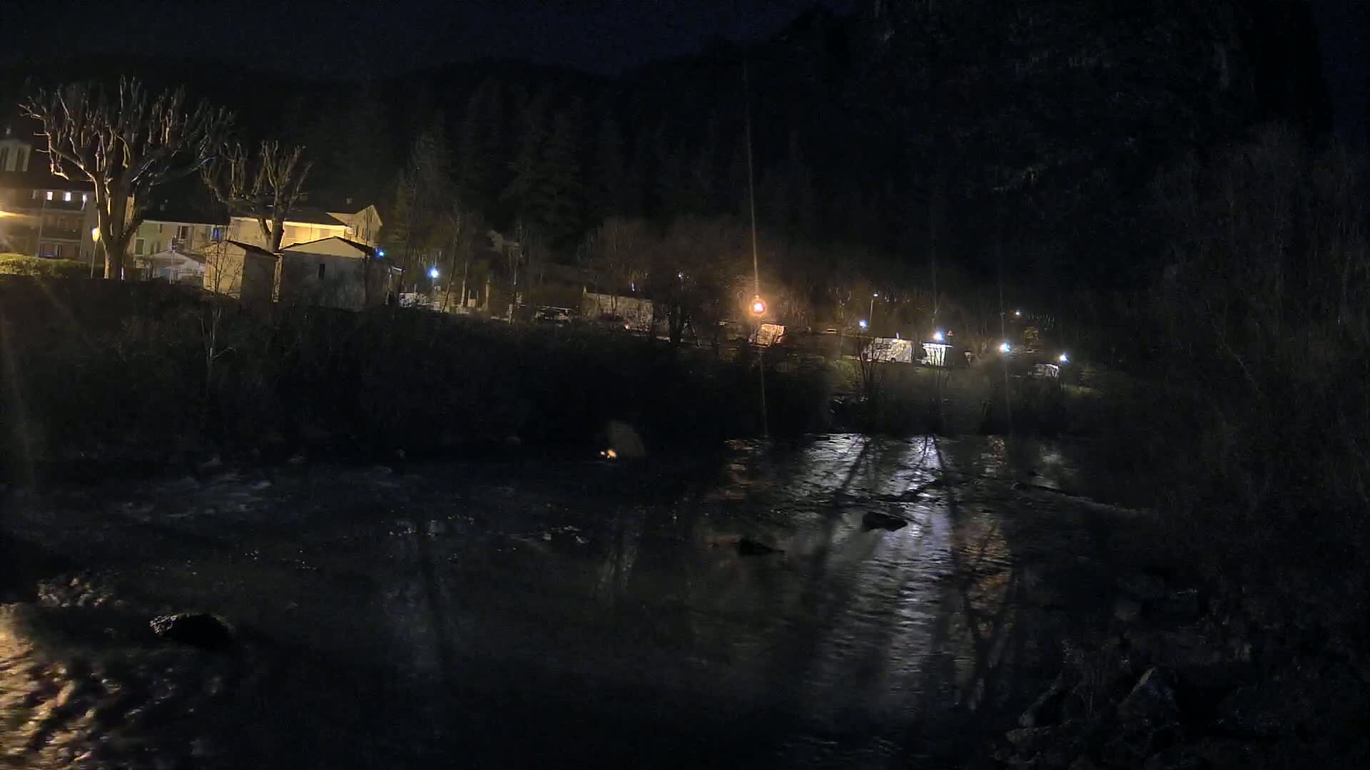 A dark river flows past a dimly lit nighttime village nestled in a hillside, under a clear, dark sky.