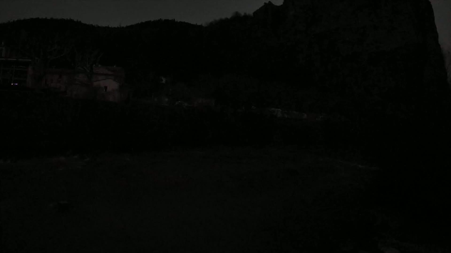 A dark, nighttime scene shows a building and trees silhouetted against a hill under a dusky sky.