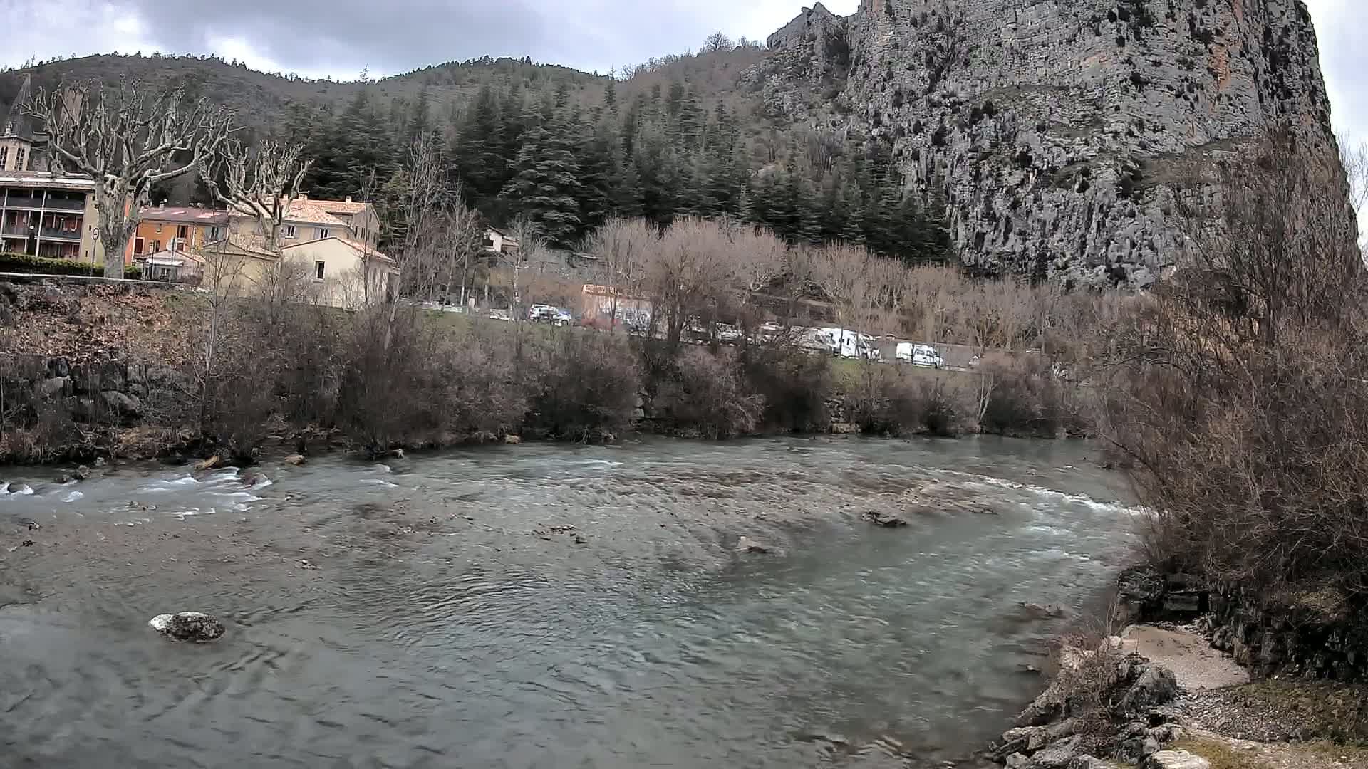 A river flows past buildings and bare trees under an overcast sky, with a large rock face rising in the background.
