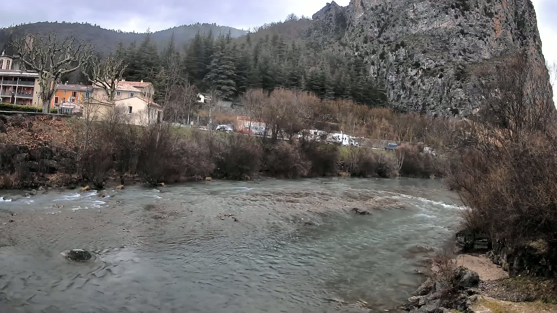 A swiftly flowing river runs past a small village nestled at the base of a large, rocky cliff under an overcast sky.