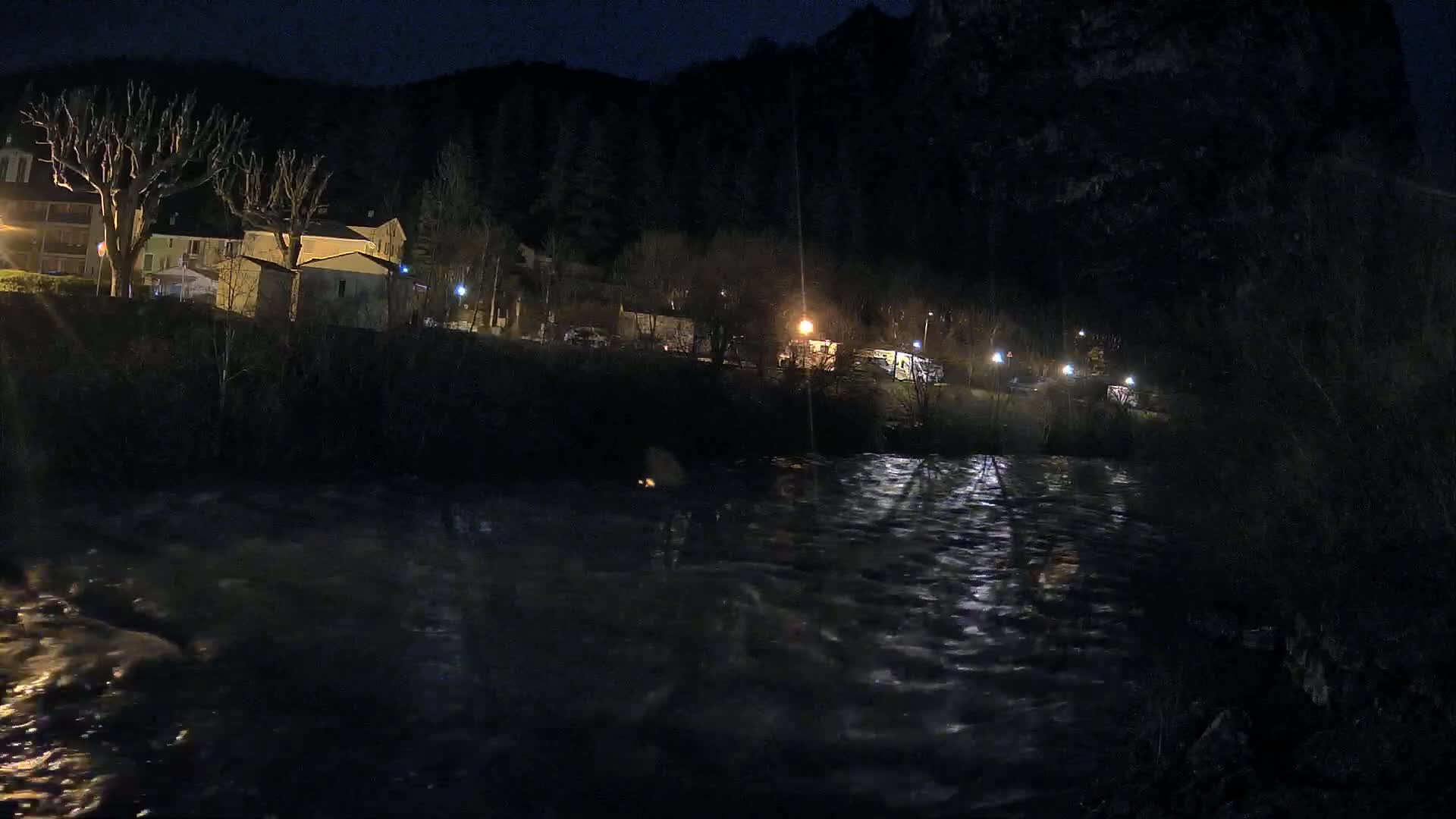 A swiftly flowing river at night runs past a small village nestled against a dark hillside under a clear, dark sky.