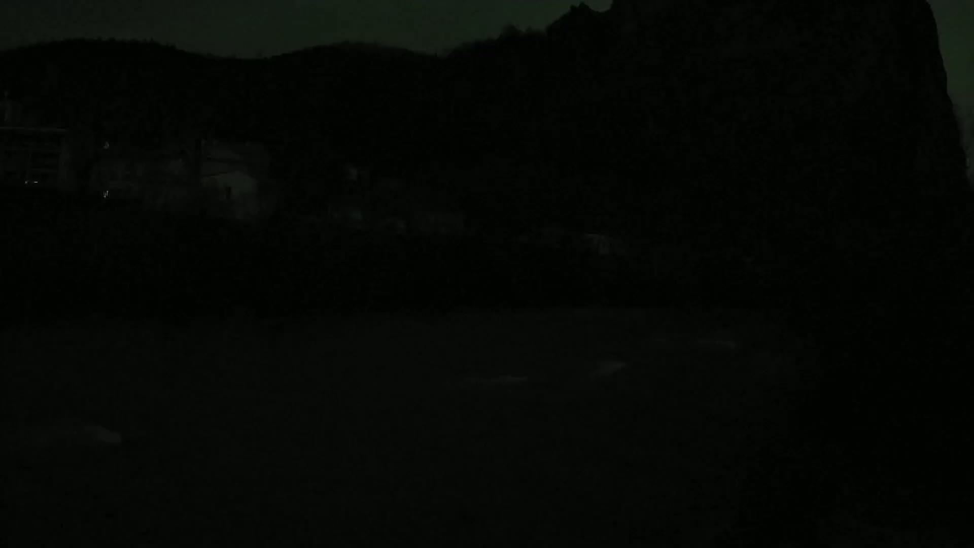 A dark, nighttime scene shows a shadowy hillside with indistinct buildings and a body of water in the foreground under a mostly clear sky.