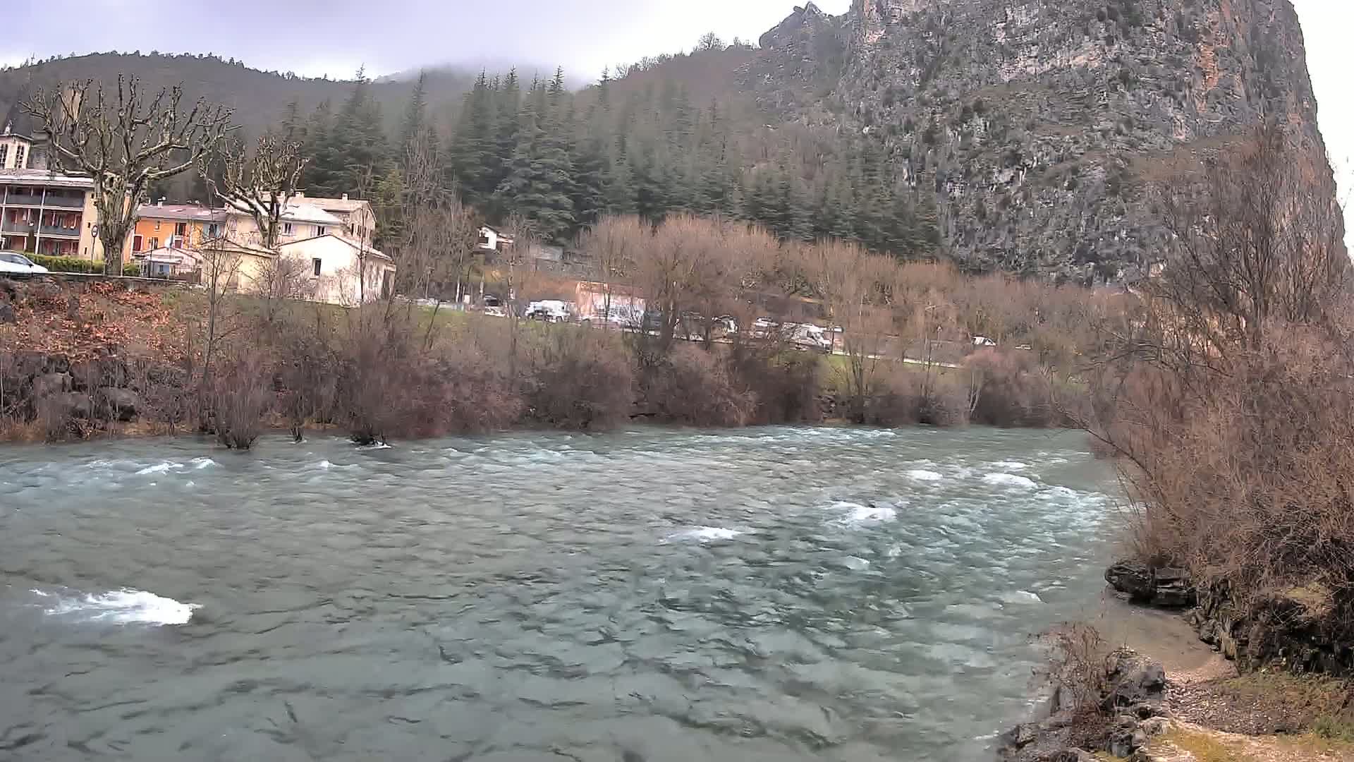 A swiftly flowing river runs in front of a village nestled at the base of a large, rocky cliff, under an overcast sky.