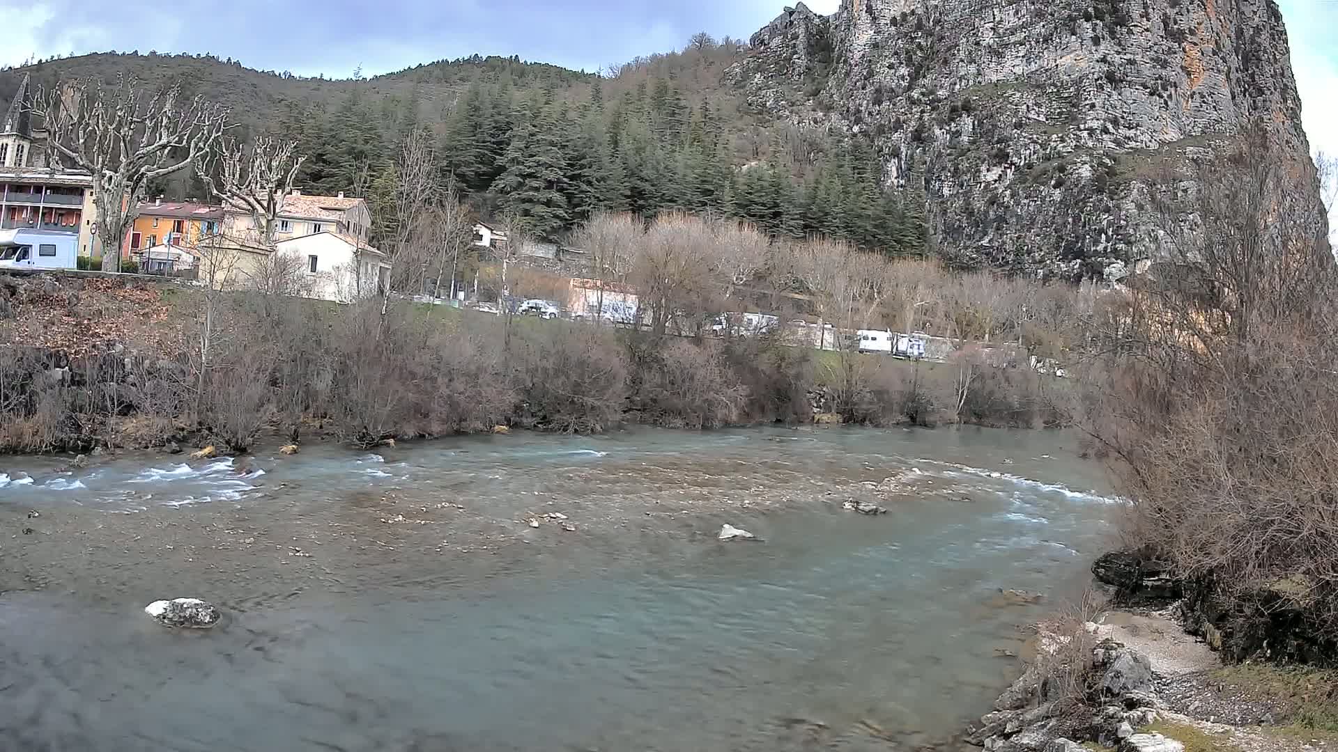 A river flows past a village nestled at the base of a rocky cliff under a cloudy sky.