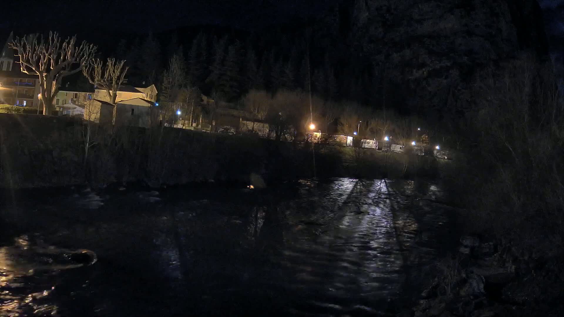 A dark, nighttime scene shows a river reflecting the lights of buildings and trees on the opposite bank, under a clear, dark sky.